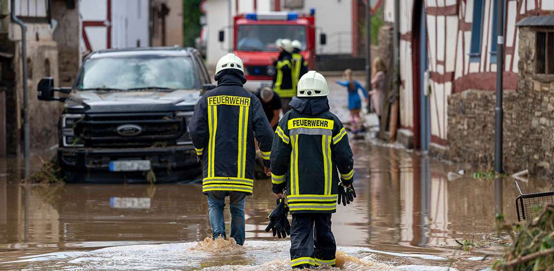 ZWei Feuerwehrleute unterwegs auf einer überschwemmten Straße. Bildquelle:  allianz-hochwasser-spende: picture alliance/dpa, Harald Tittel