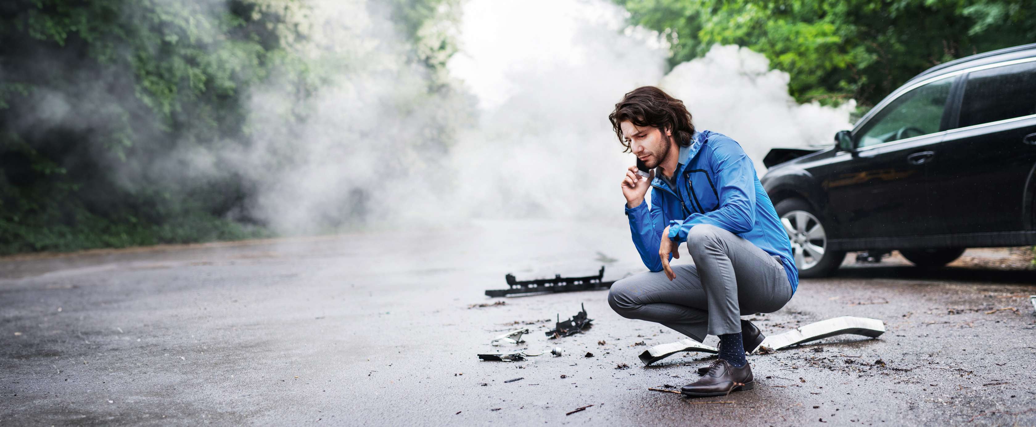 Allianz  - Kfz-Versicherung Fahrerschutzversicherung Angebot: Allianz  - Kfz-Versicherung, Auslandsschadenschutz Angebot: Ein Mann in blauer Jacke hockt nachdenklich am Straßenrand. Im Hintergrund ist Rauch zu sehen, auf der Straße liegen Autoteile.