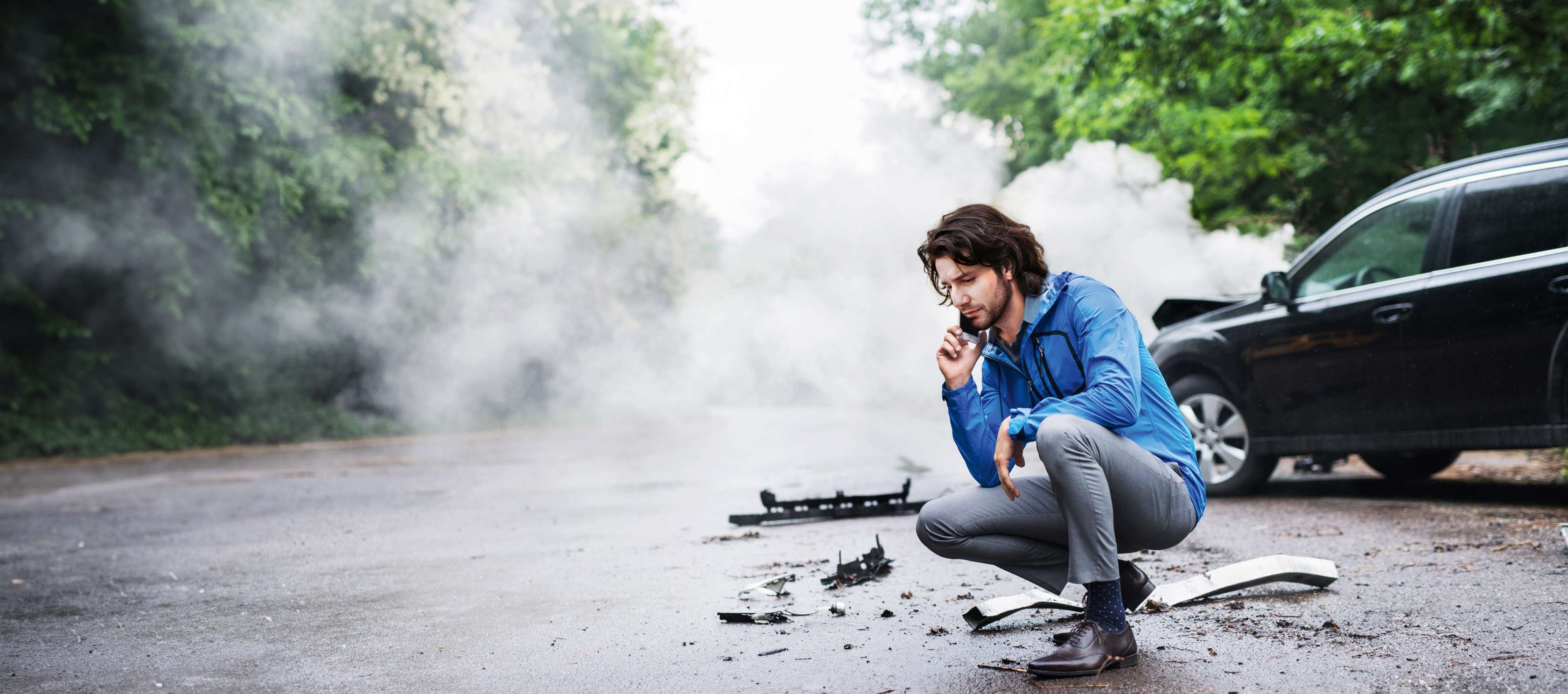 GAP-Versicherung: Mann kniet telefonierend auf Landstraße vor Auto mit Frontschaden