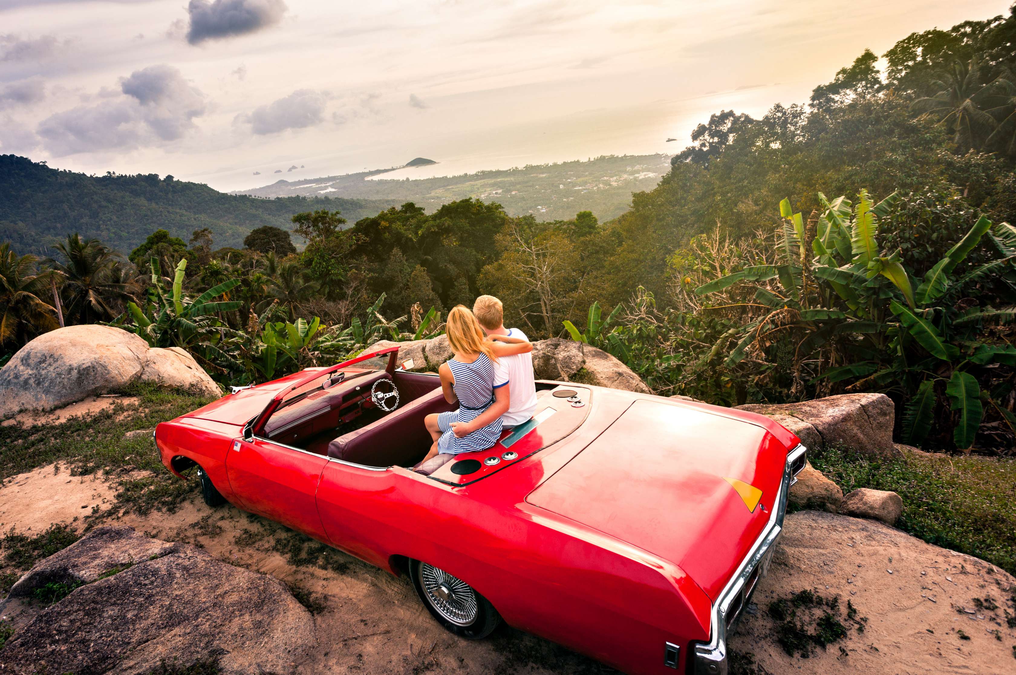 Allianz - Oldtimer Versicherung Angebot: Rotes Oldtimer-Cabrio mit offenem Verdeck und einem jungen Pärchen auf der Rückbank parkt am Rand einer Panorama-Bergstraße mit Blick über grüne Täler.