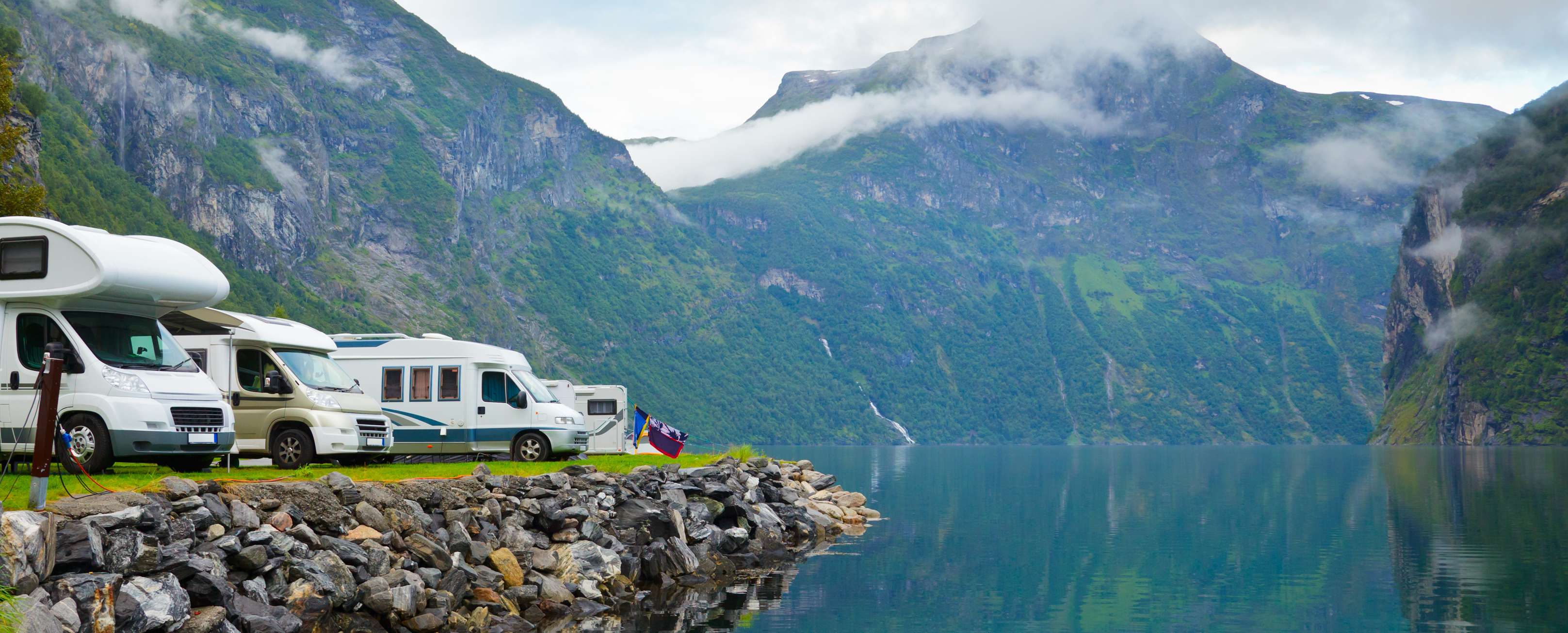 Allianz - Wohnmobilversicherung Angebot: Wohnmobil Versicherung: Mehrere Campingfahrzeuge stehen an Ufer eines Bergsees.