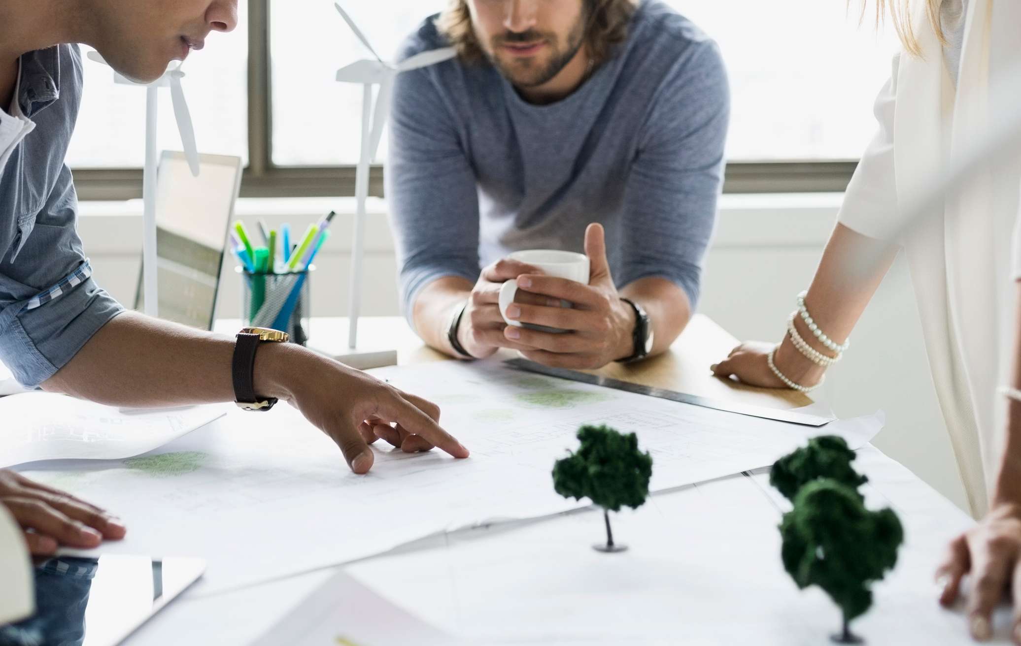 Allianz Berufshaftpflichtversicherung Angebot: Allianz Berufshaftpflichtversicherung: Drei Personen stehen um einen Tisch und diskutieren über Architekturpläne. Zu sehen sind ihre Unterarme und Hände. Die linke Person trägt ein Jeanshemd und deutet mit dem Finger auf einen bestimmten Punkt in einem Plan , die mittlere hält eine Tasse in den Hönden und die rechte trägt mehrere Perlenarmbänder.