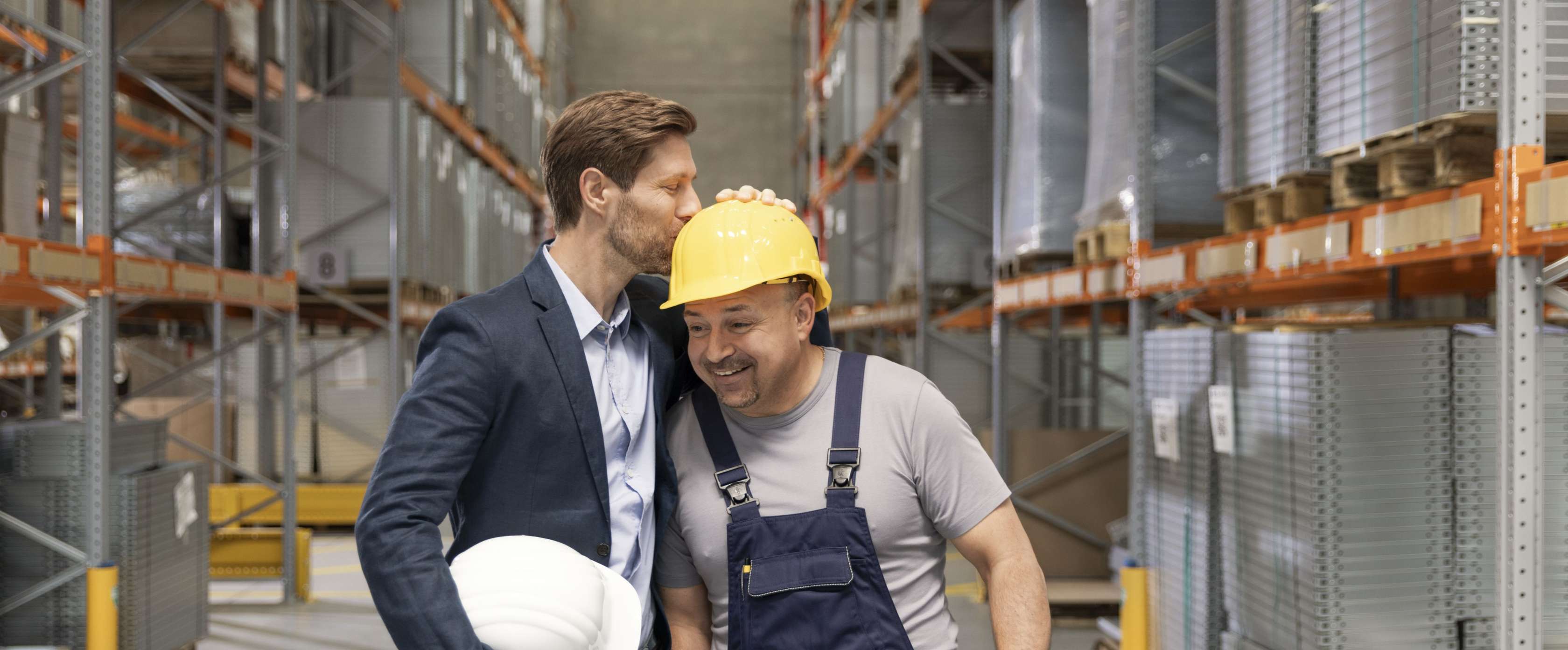 Allianz - Betriebliche Vorsorge im Unternehmen Angebot: Der im Anzug gekleidete Chef einer Baufirma küsst einen seiner Mitarbeiter auf seinen gelben Helm.