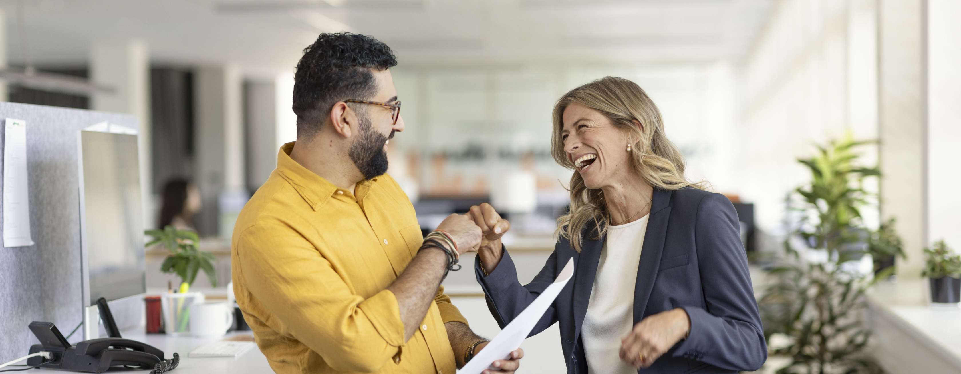 Allianz - Betriebliche Altersversorgung für Unternehmen Angebot:  Zwei Personen, ein Mann  im gelben Hemd und Brille sowie eine Frau mit langen blonden Haaren in einem dunkelblauen Blazer, lachen und geben sich die Faust.