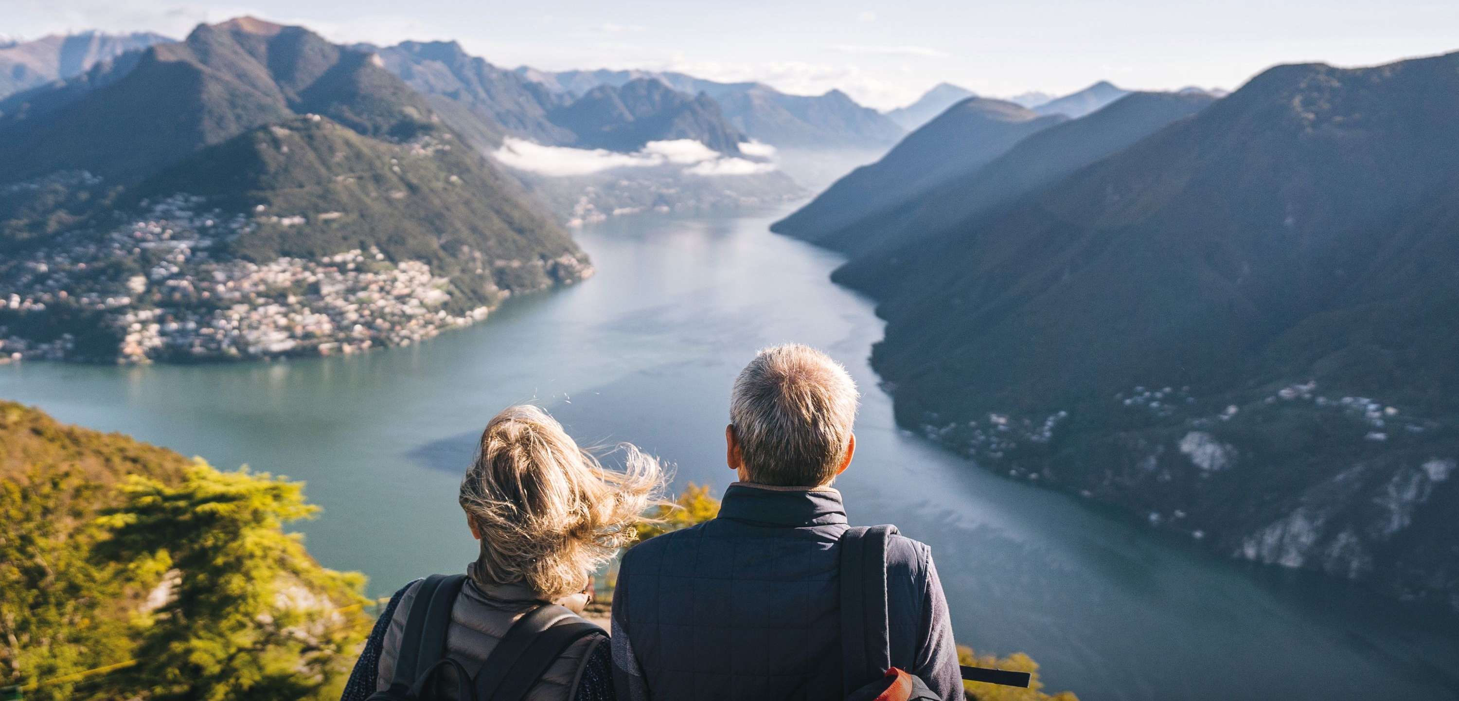 Ein Paar sitzt auf einer Aussichtsplattform und blickt auf einen See, der sich zwischen hohen Bergen erstreckt. Im Hintergrund sind eine Stadt und schneebedeckte Gipfel zu sehen