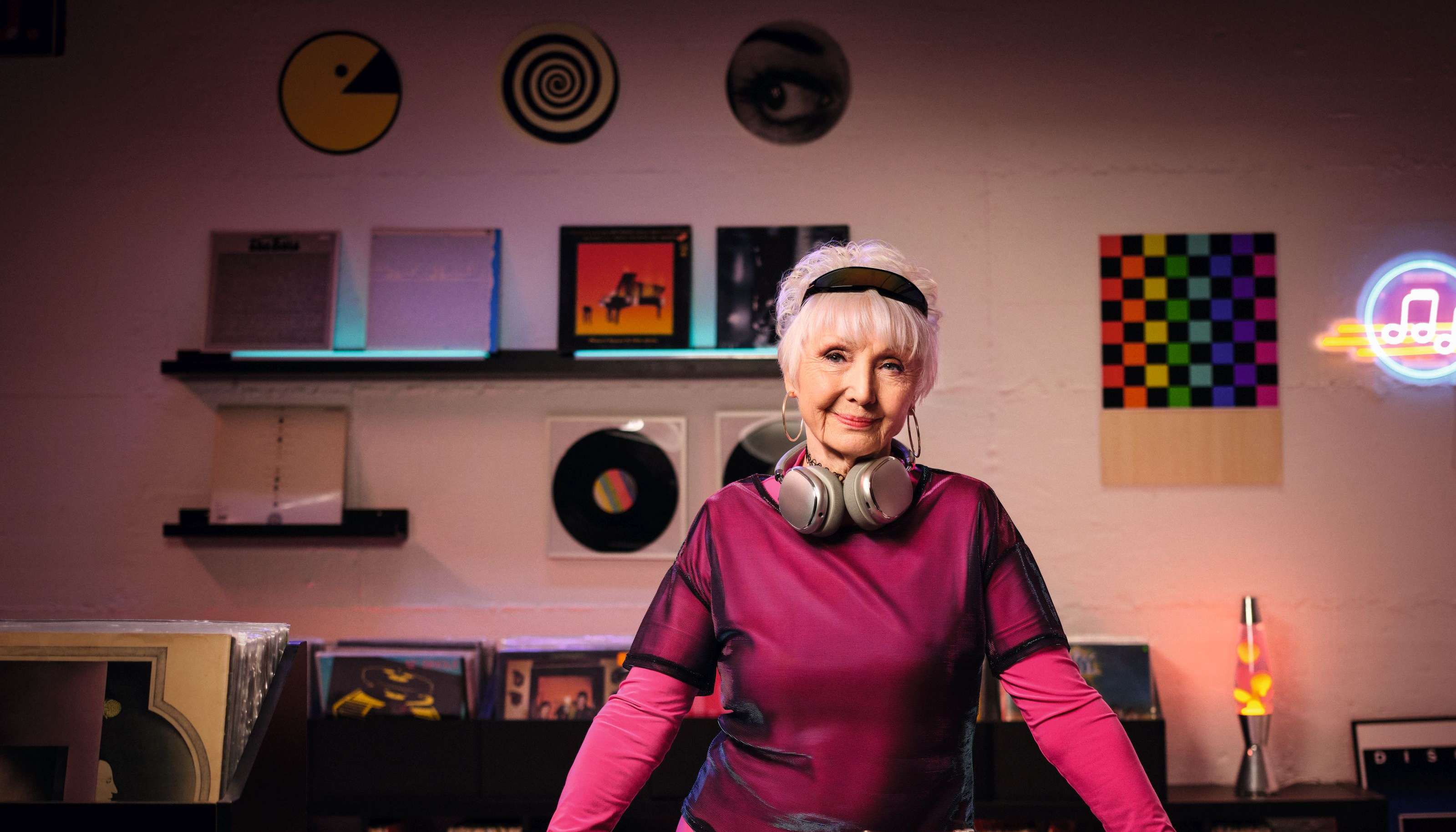 Allianz Pflegezusatzversicherung: Eine ältere Dame in pinker Kleidung steht hinter einem DJ-Pult