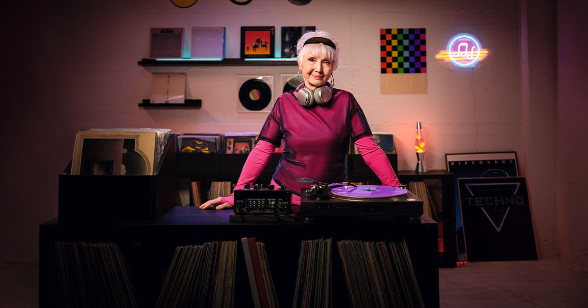 Allianz Pflegezusatzversicherung: Eine ältere Dame in pinker Kleidung steht hinter einem DJ-Pult