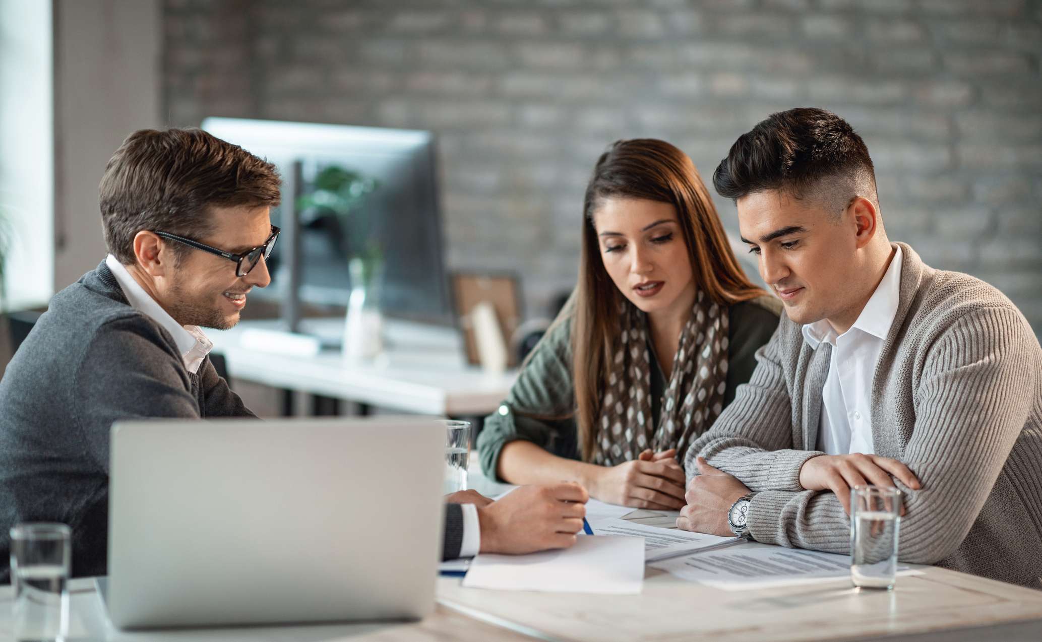  Allianz - Angebote für Kunden der Commerzbank: Drei Personen sitzen an einem Tisch in einem modernen Büro. Sie sind in ein Gespräch vertieft und schauen auf Dokumente, die auf dem Tisch liegen. Auf dem Tisch stehen auch zwei Gläser Wasser und ein Laptop.