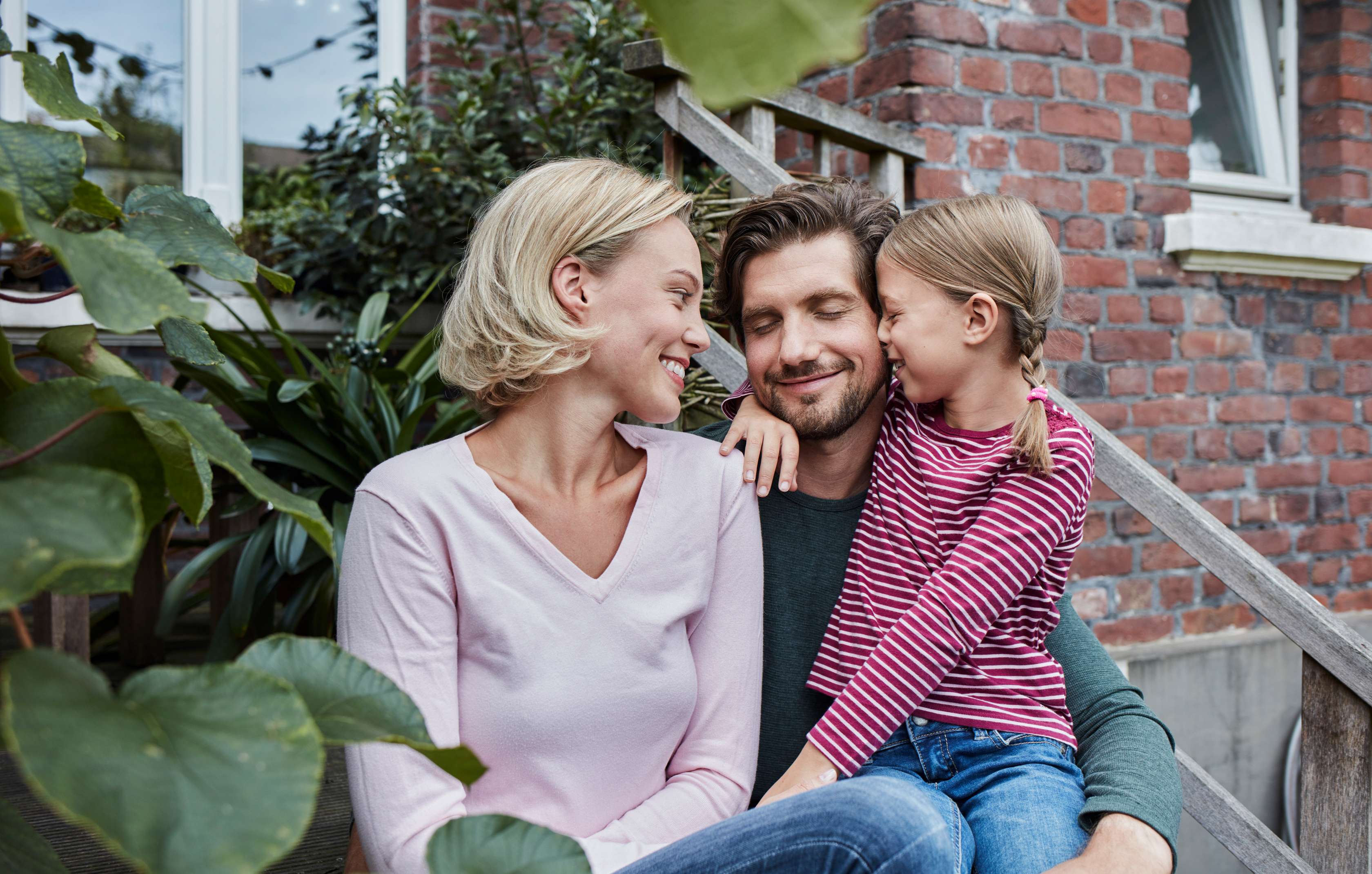 Allianz Kooperation - Risiko­lebens­versicherung (RLV):  Vater, Mutter und Tochter kuscheln vor einem Haus.