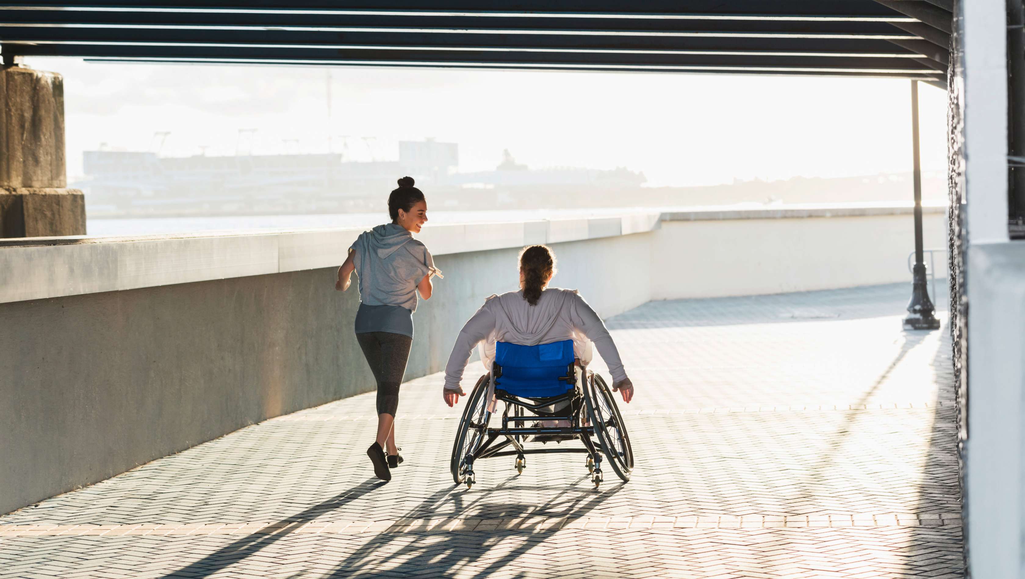 Allianz - Unfallversicherung Angebot: Ein Rollstuhlfahrer fährt neben einer Joggerin auf einem Weg.