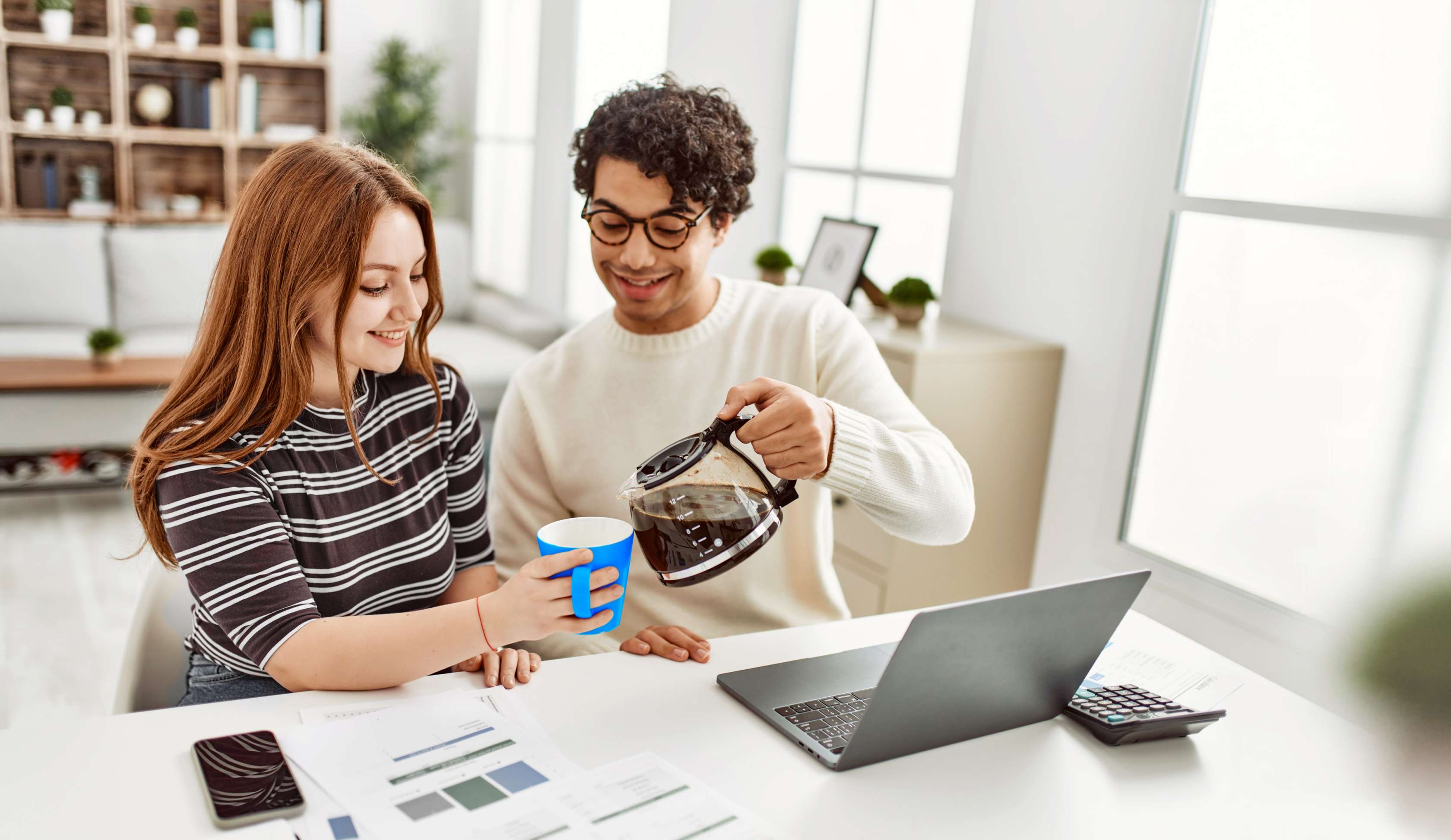 Allianz Privathaftpflicht: Ein junger Mann sitzt neben einer jungen Frau an einem Laptop, während er ihr Kaffee in eine Tasse schenkt