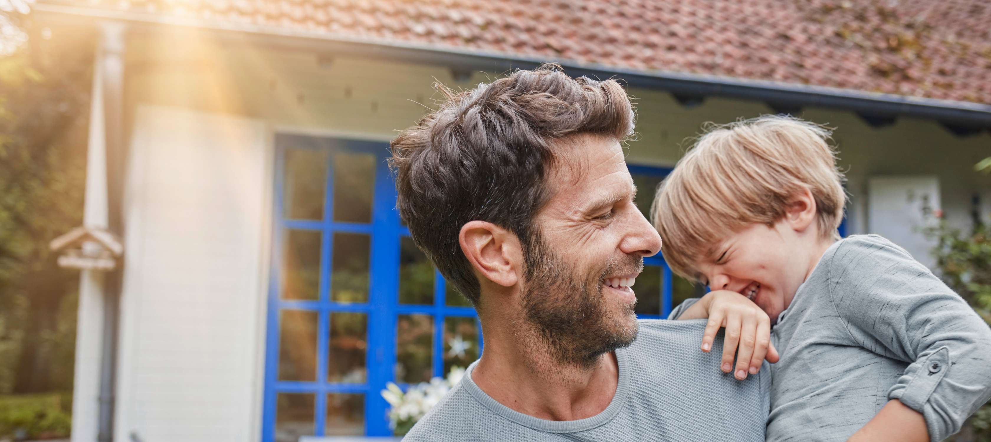 Wohngebäudeversicherung: Vater steht mit Sohn auf dem Arm glücklich vor seinem Haus