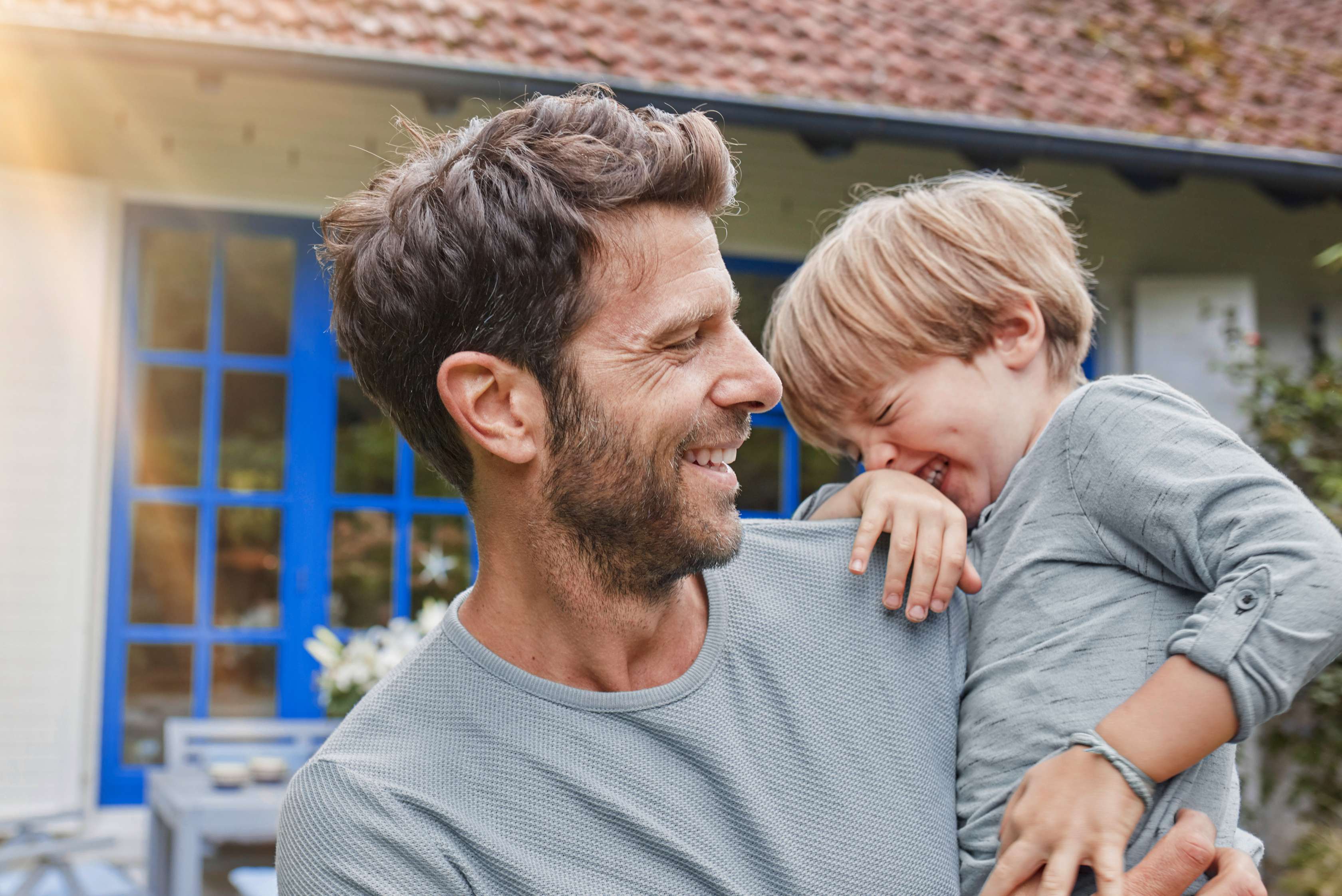 Wohngebäudeversicherung: Vater steht mit Sohn auf dem Arm glücklich vor seinem Haus