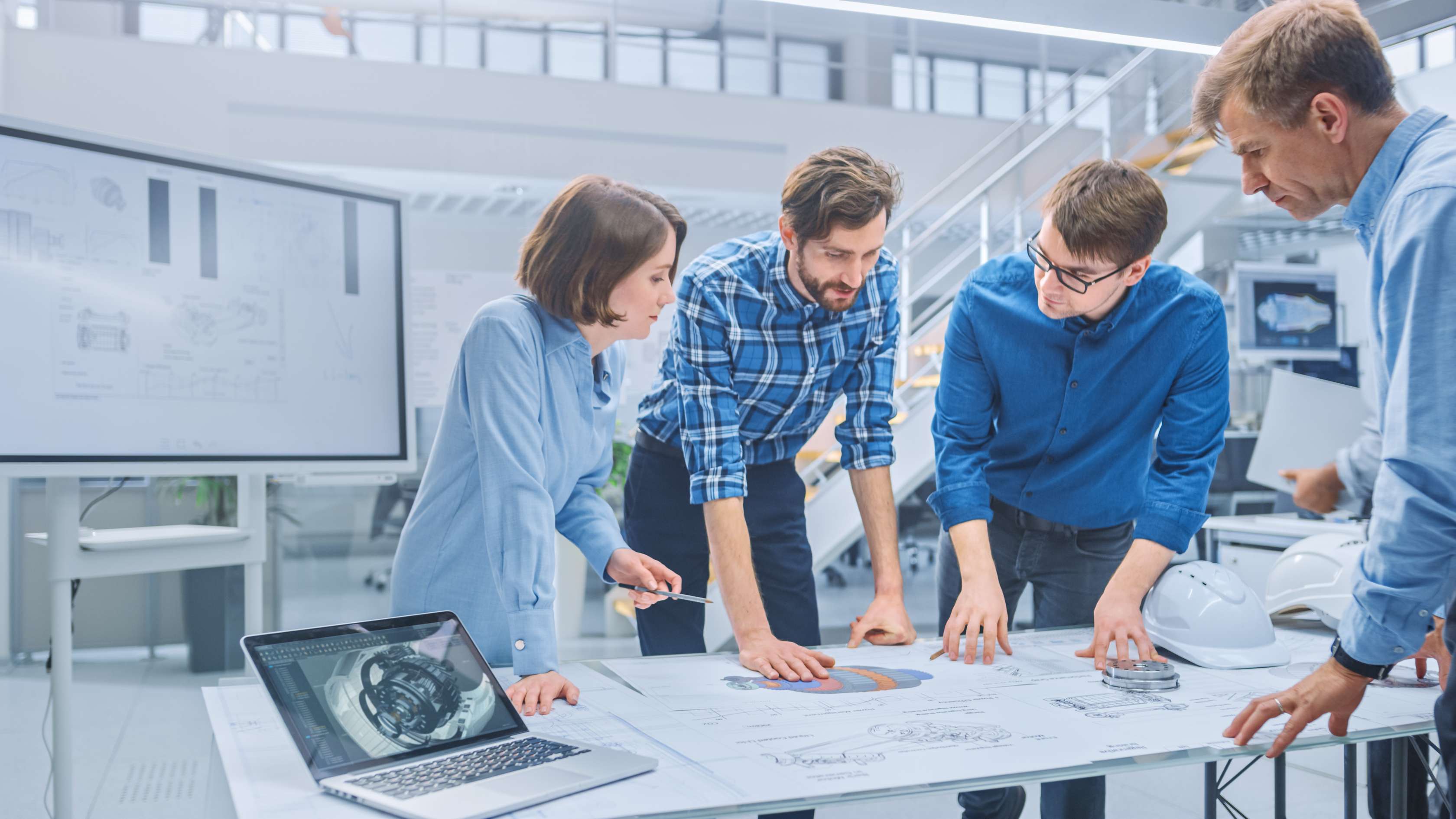 Allianz - Betriebshaftpflichtversicherung: Vier Ingenieure und Ingenieurinnen stehen gemeinsam an einem Schreibtisch und diskutieren über die vor ihnen liegenden Pläne und Zeichnungen.