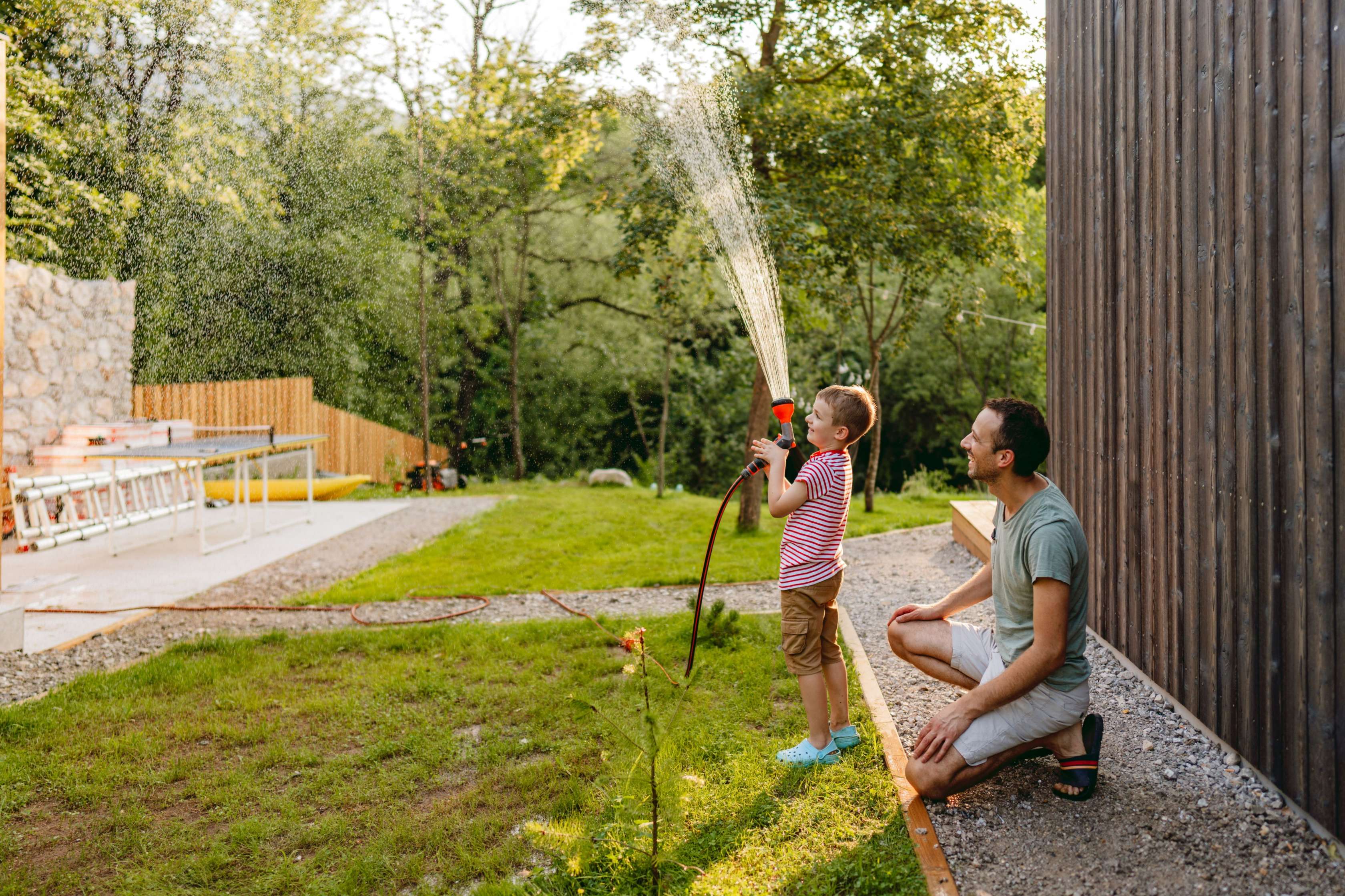 KinderPolice Kooperationsangebot: Ein Vater steht im Garten mit seinem kleinen Sohn und sie gießen gemeinsam mit einem Schlauch den Garten.