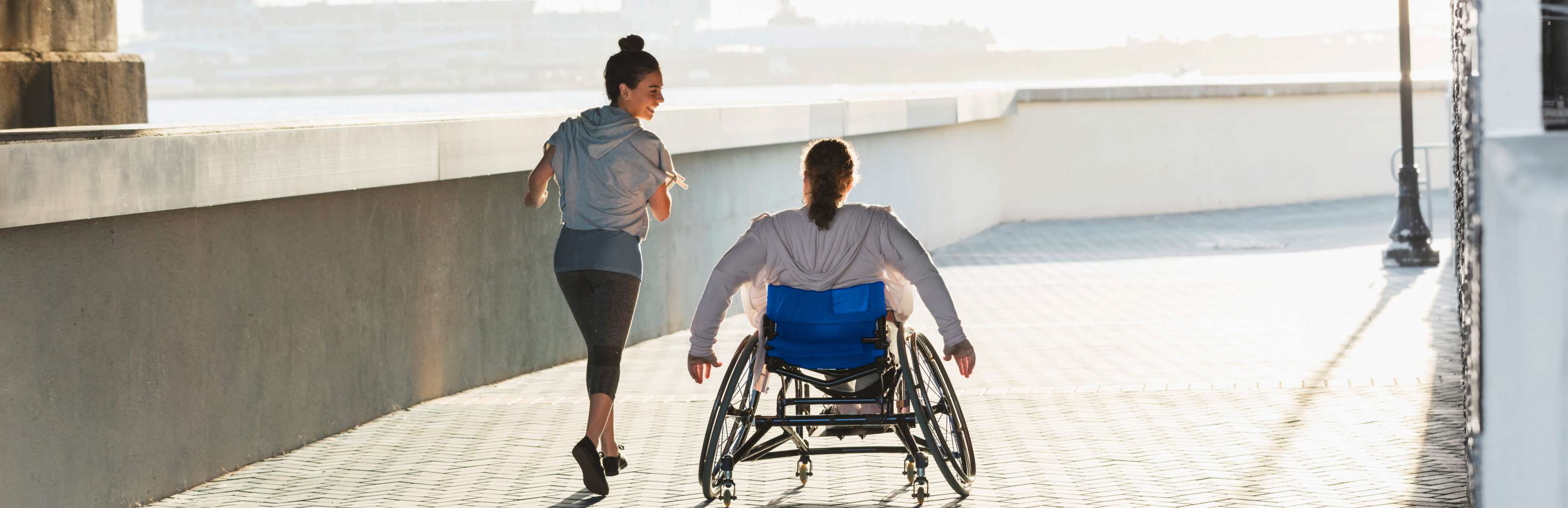 Allianz Unfallversicherung: Ein Rollstuhlfahrer fährt neben einer Joggerin auf einem Weg