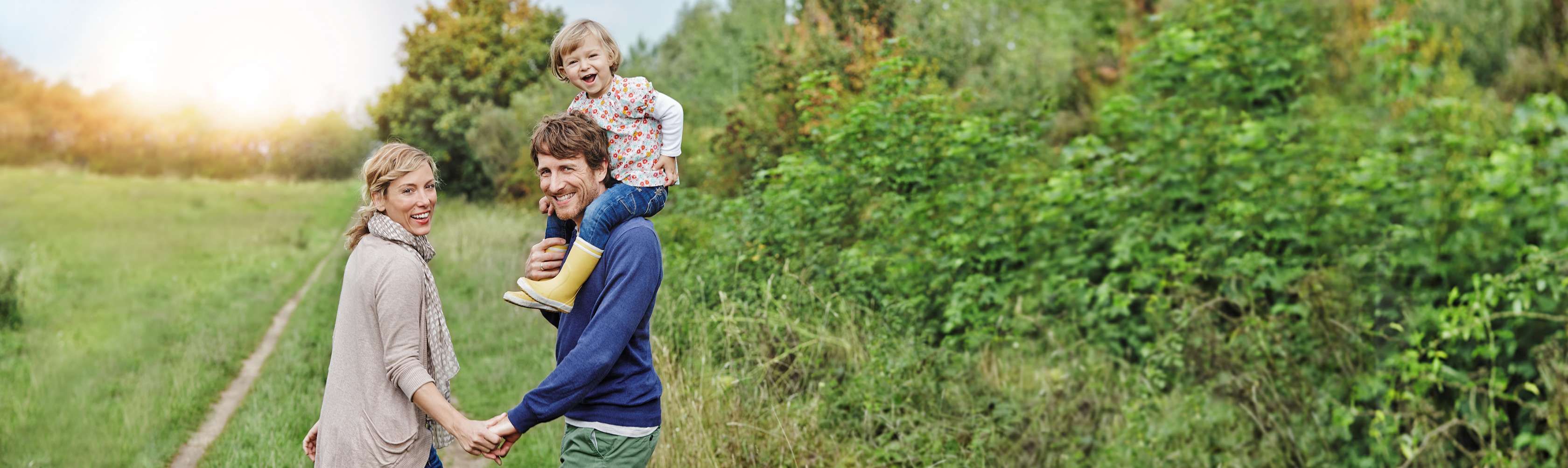 Frau und Mann mit Kind auf den Schultern spazieren lachend über einen Feldweg