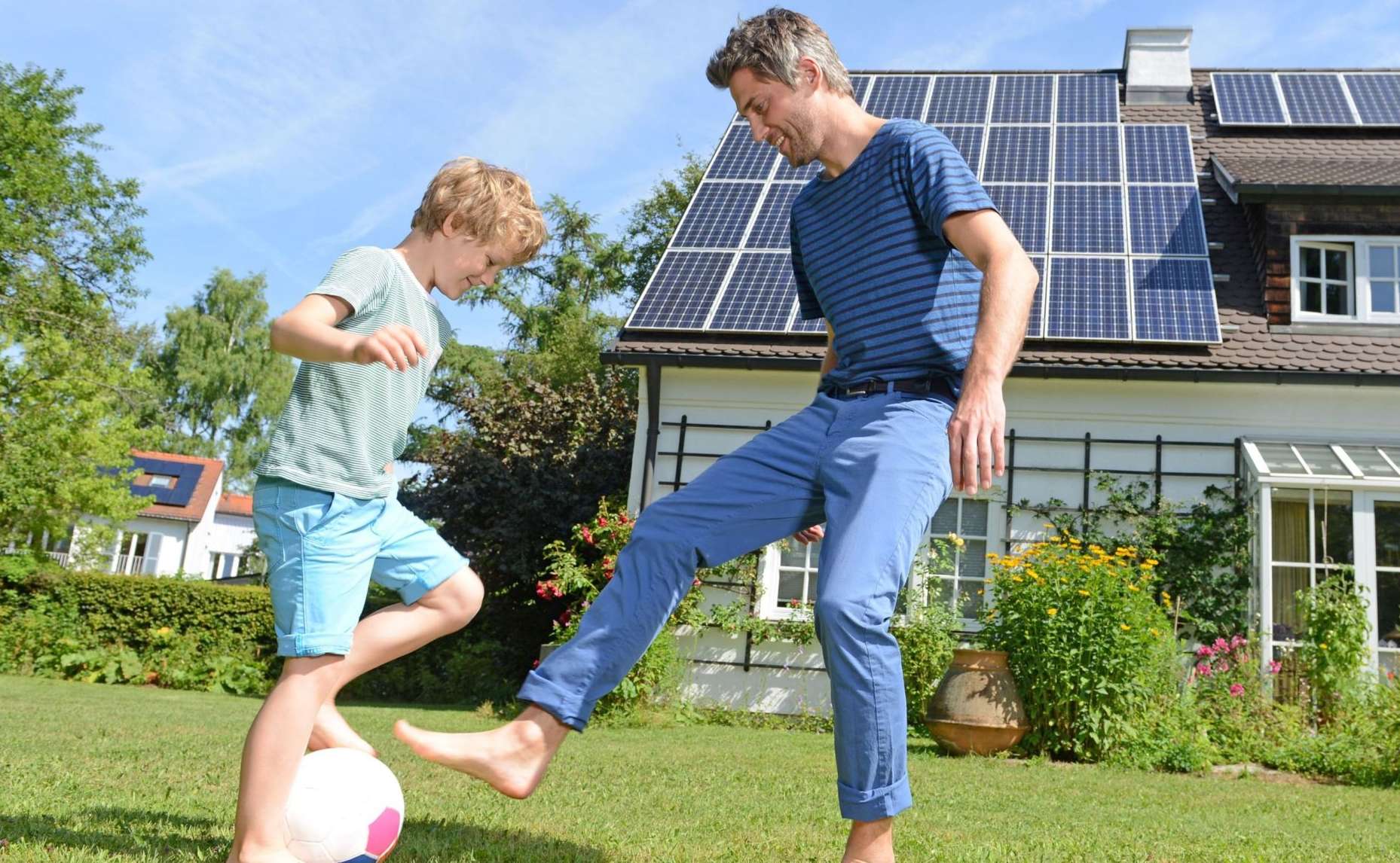 Allianz - Baufinanzierung: Ein Erwachsener und ein Kind spielen barfuß Fußball auf einer grünen Wiese vor einem Haus mit Solarpaneelen auf dem Dach.