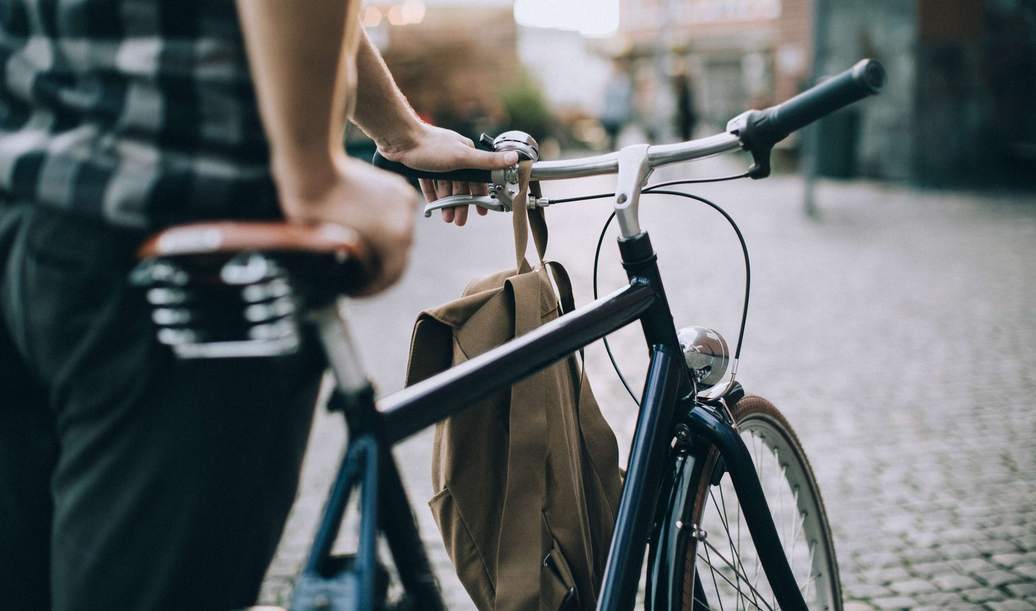 Eine Person in einem karierten Hemd hält den Lenker eines Fahrrads. Am Rahmen des Fahrrads ist eine braune Tasche befestigt. Im Hintergrund ist eine gepflasterte Straße und unscharfe Gebäude zu sehen.