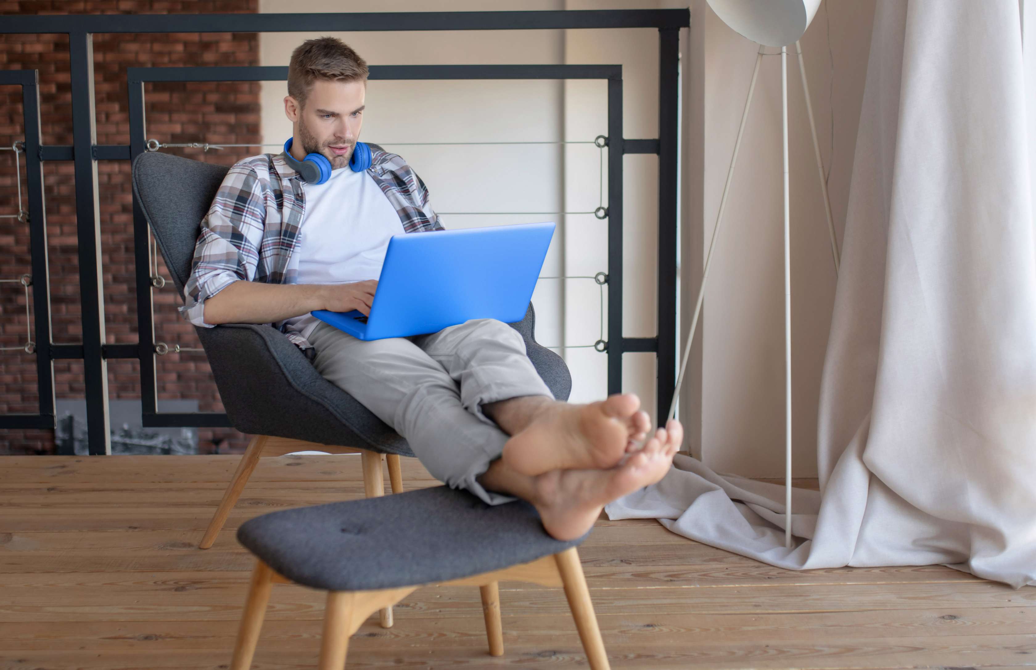 Allianz - Gegenstandsschutz Laptop: Eine Person sitzt in einem modernen Wohnzimmer auf einem grauen Sessel und arbeitet an einem blauen Laptop. Die Person hat die Beine auf einen Hocker gelegt und trägt eine karierte Hemdjacke sowie eine helle Hose. Im Hintergrund ist eine Backsteinwand zu sehen, daneben steht eine weiße Stehlampe vor bodenlangen Vorhängen.