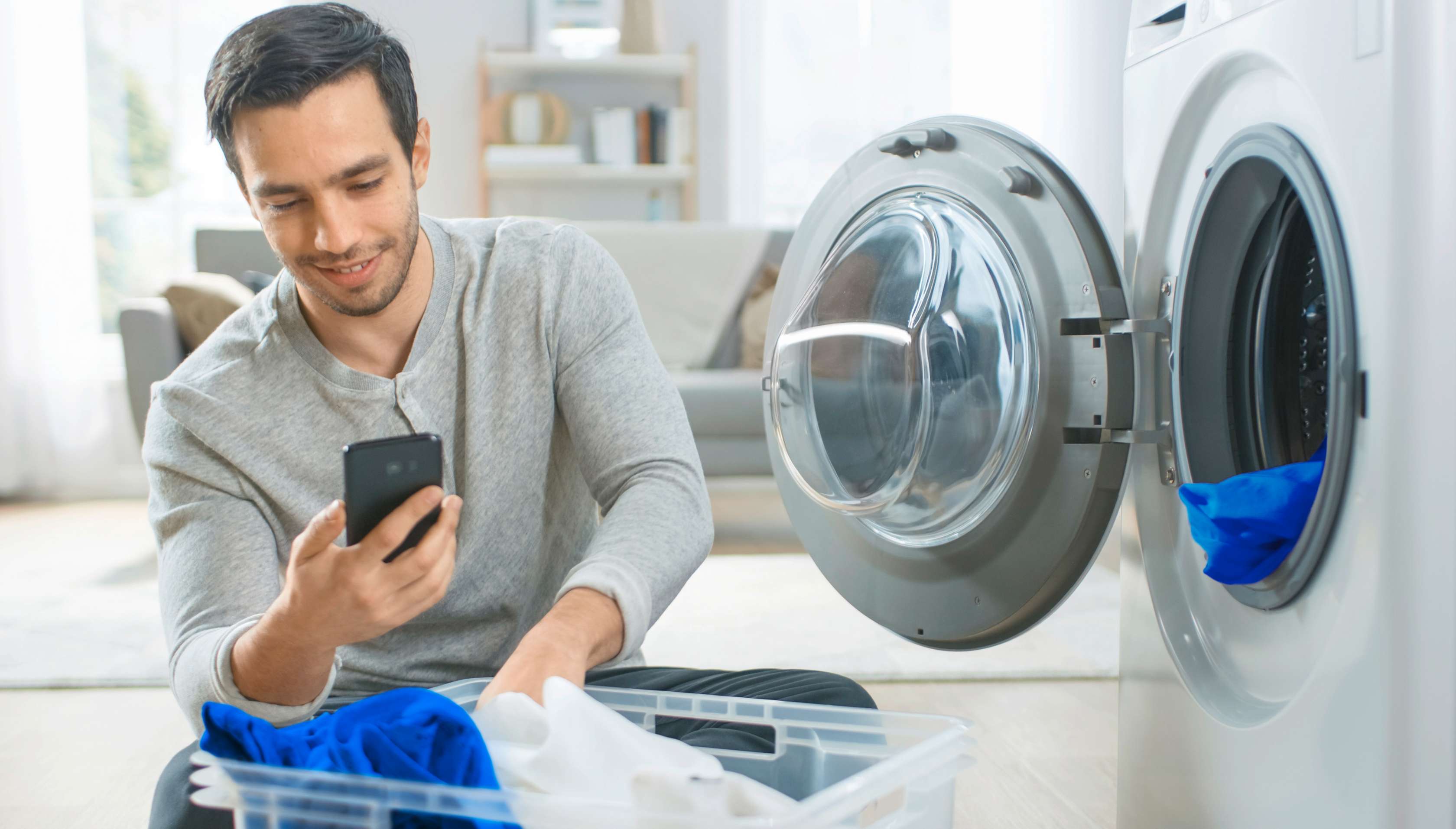 Allianz Hausrat-Haftpflicht-Kombi. Mann kniet vor einer Waschmaschine und schaut lächelnd auf sein Handy.