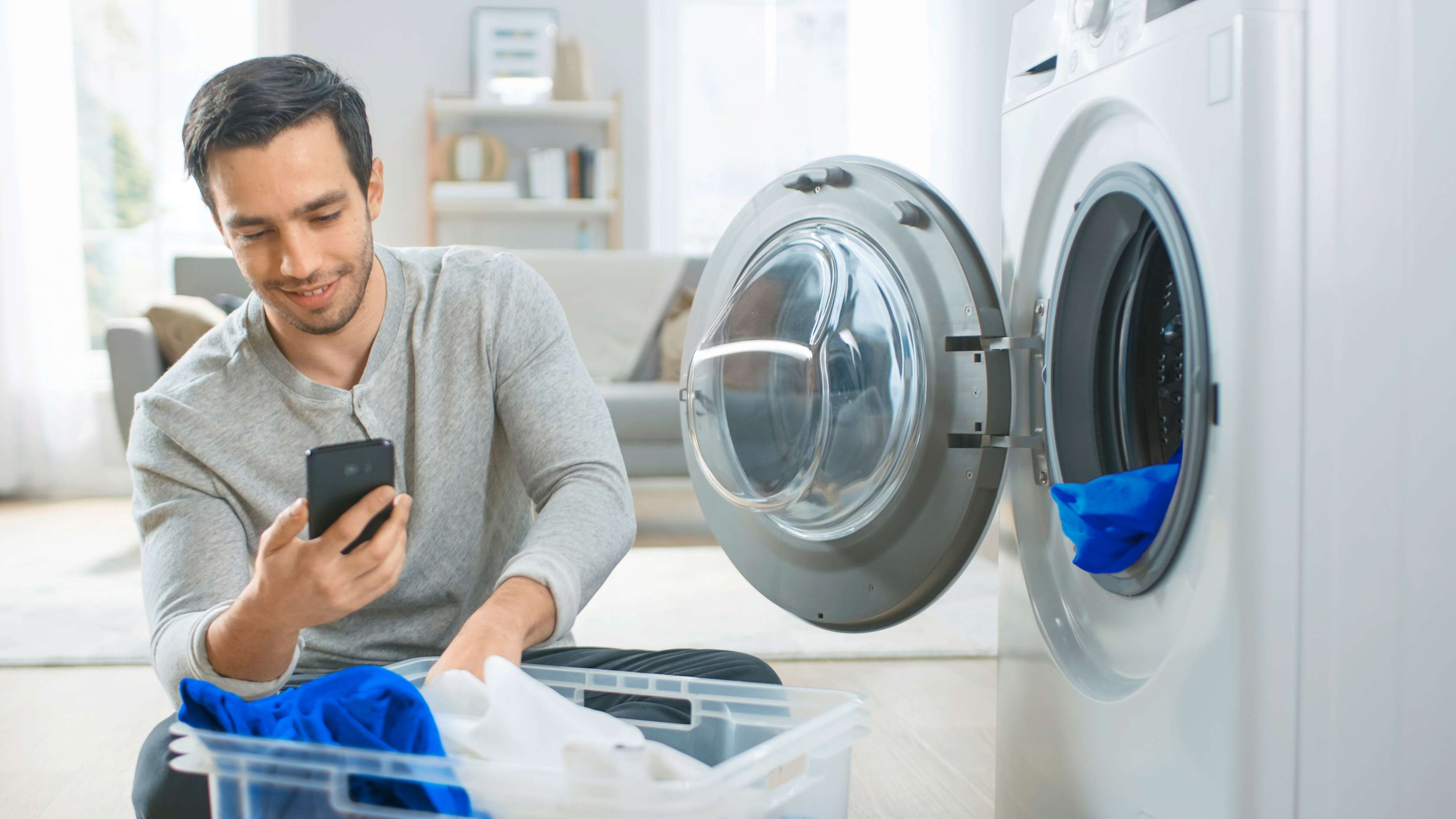 Allianz Hausrat-Haftpflicht--Rechtsschutz-Kombi: Mann kniet vor einer Waschmaschine und schaut lächelnd auf sein Handy.