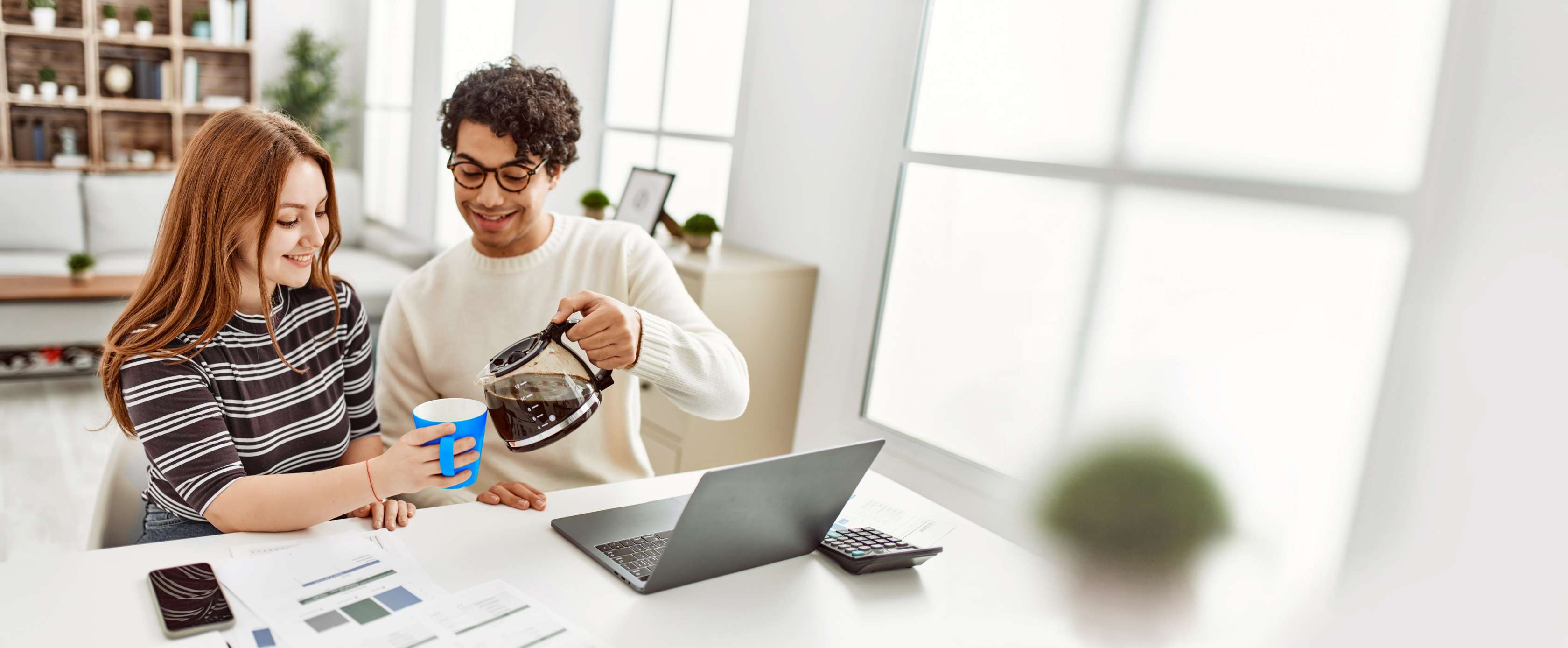 Allianz Privathaftpflicht: Ein junger Mann sitzt neben einer jungen Frau an einem Laptop, während er ihr Kaffee in eine Tasse schenkt