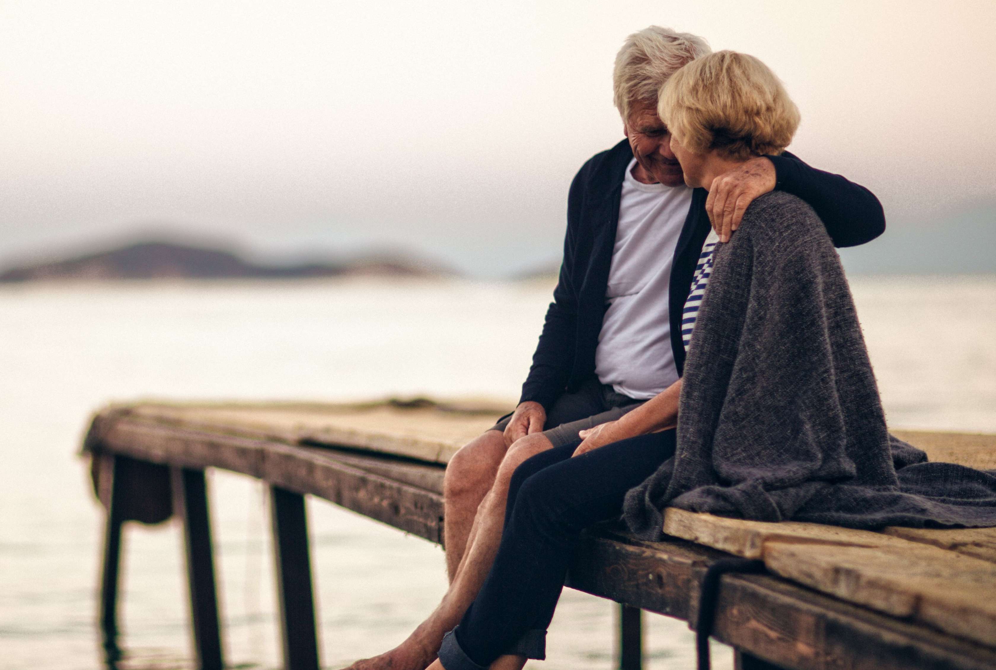 Allianz Rechtschutzversicherung Berliner Testament: Älteres Paar sitzt Arm in Arm auf einem Steg am Wasser.