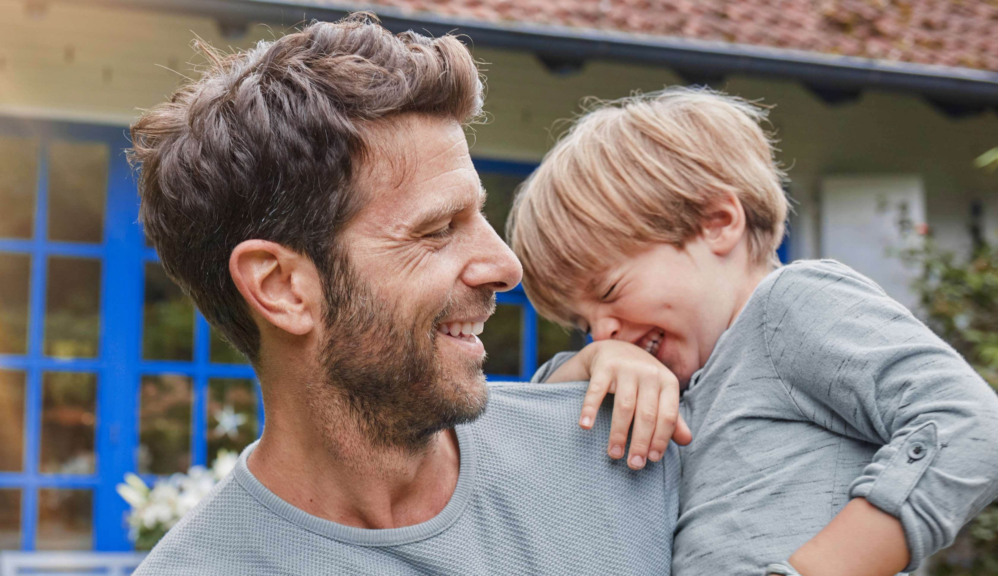 Allianz Wohngebäudeversicherung: Vater lacht mit seinem Sohn auf dem Arm vor einem Haus.