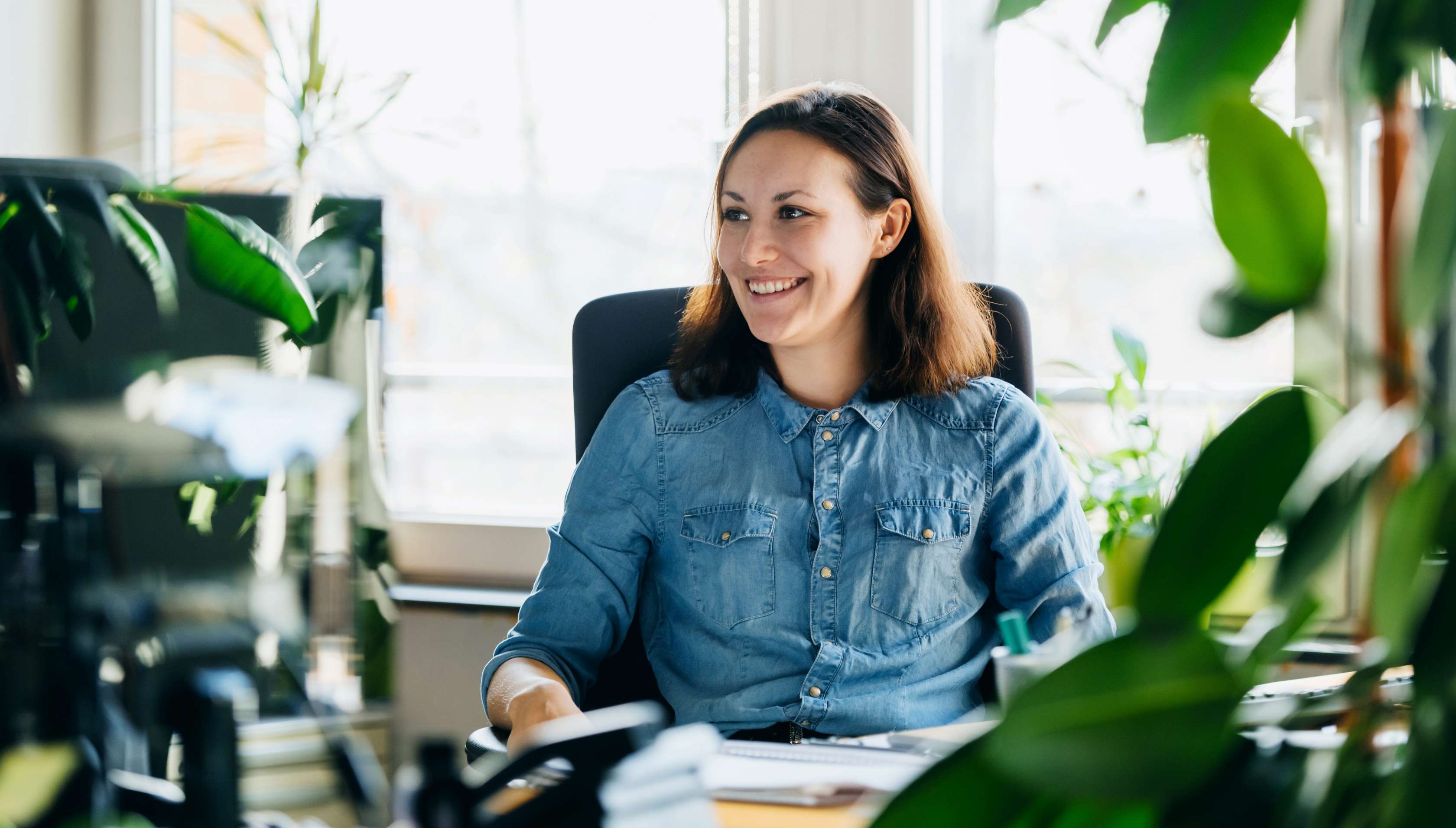 Allianz - Versicherungen für Beamte Angebot: Das Bild zeigt eine fröhliche Frau am Schreibtisch im Büro umgeben von vielen Zimmerpflanzen.