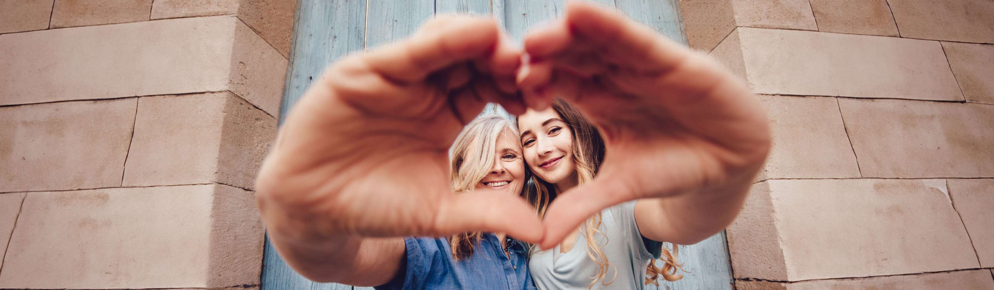 Sterbegeldversicherung – Mutter und Tochter lächeln in die Kamera und bilden mit ihren Händen zusammen ein Herz.