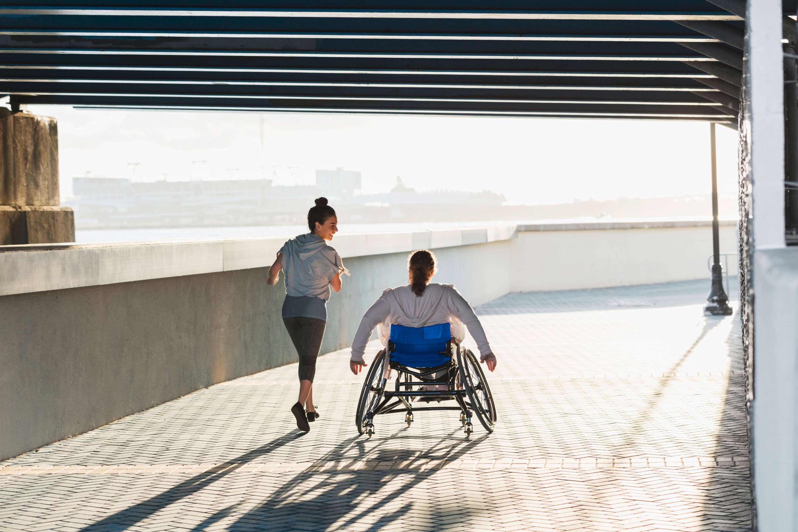 Allianz Unfallversicherung: Ein Rollstuhlfahrer fährt neben einer Joggerin auf einem Weg