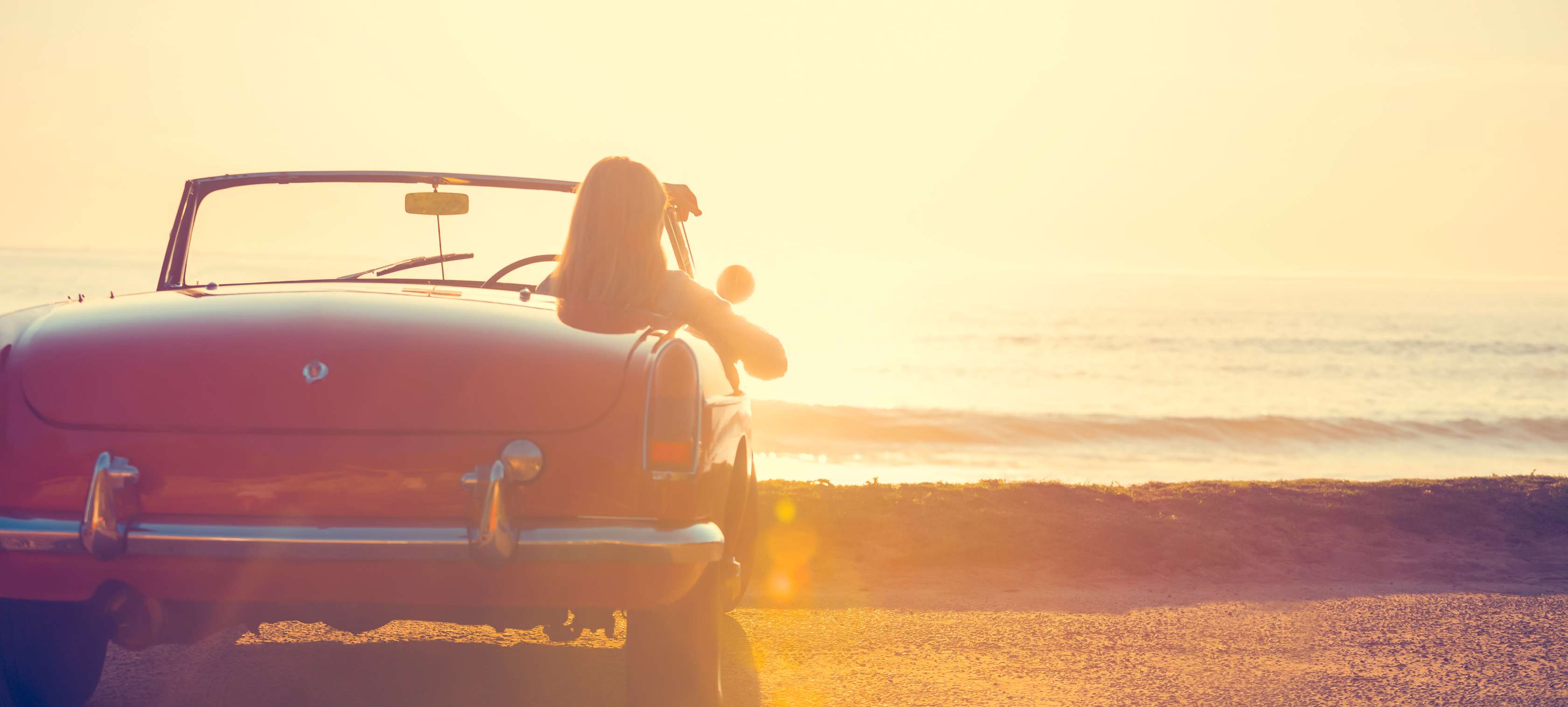 Frau sitzt am Steuer eines Oldtimer-Cabrios und genießt Sonnenuntergang am Strand