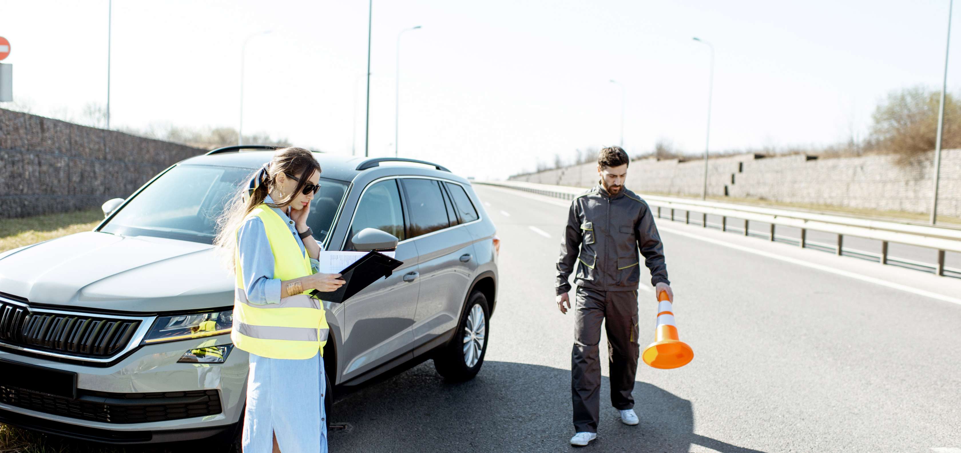 Autounfall: Ein Auto steht am Straßenrand, Frau hält Papiere in der Hand, ein Mann stellt ein Warnhütchen auf