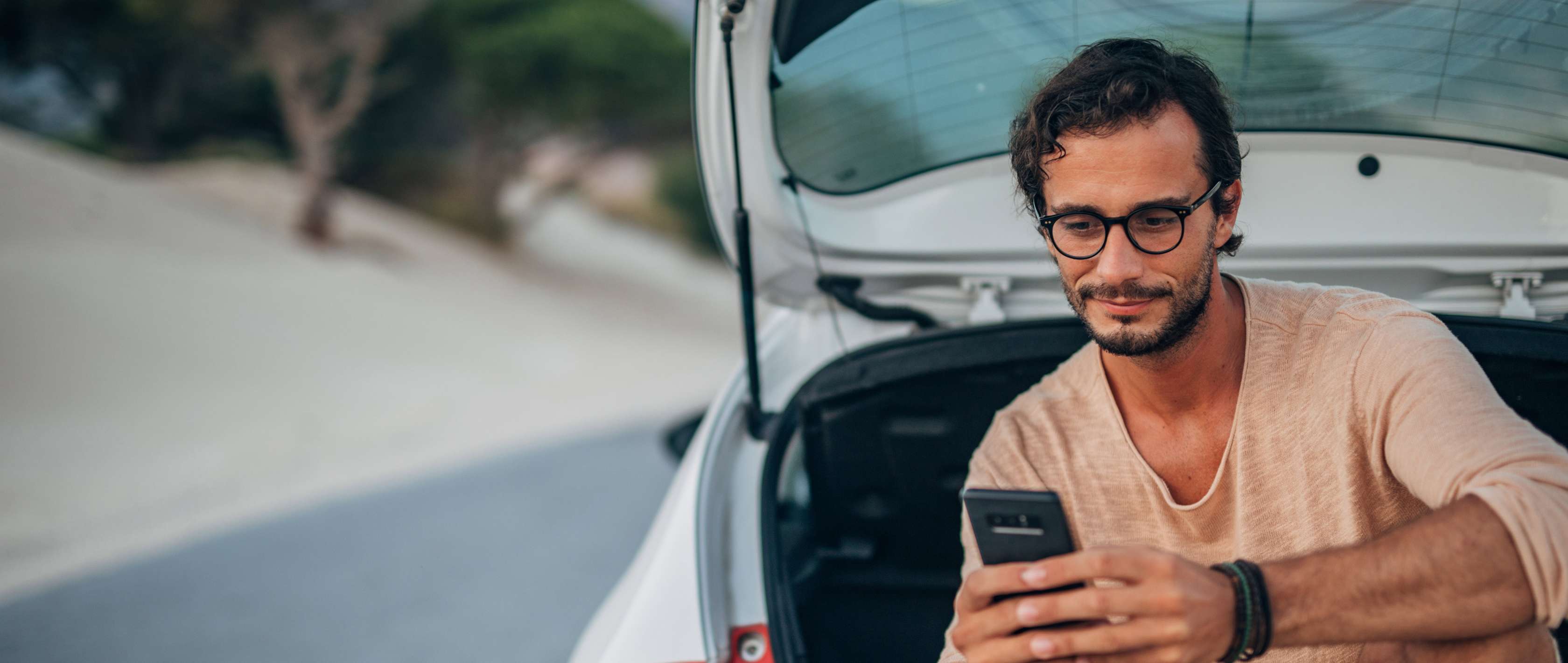 Einstufung Kfz-Versicherung: Mann sitzt in Kofferaum eines Pkw und blickt auf Smartphone