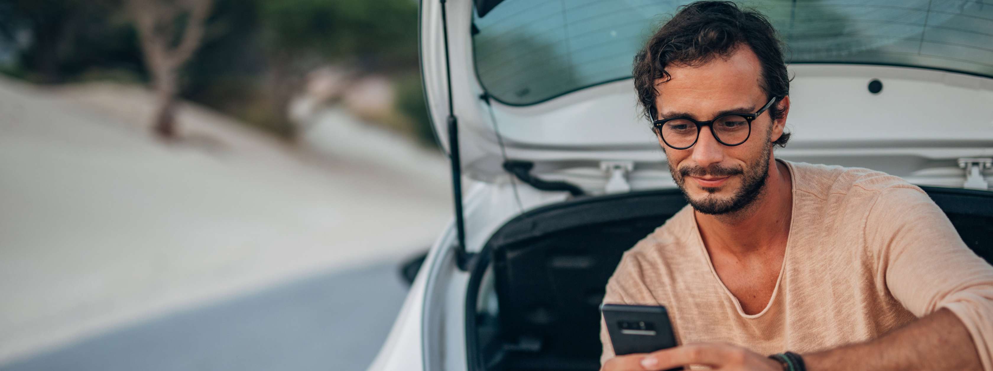 Einstufung Kfz-Versicherung: Mann sitzt in Kofferaum eines Pkw und blickt auf Smartphone