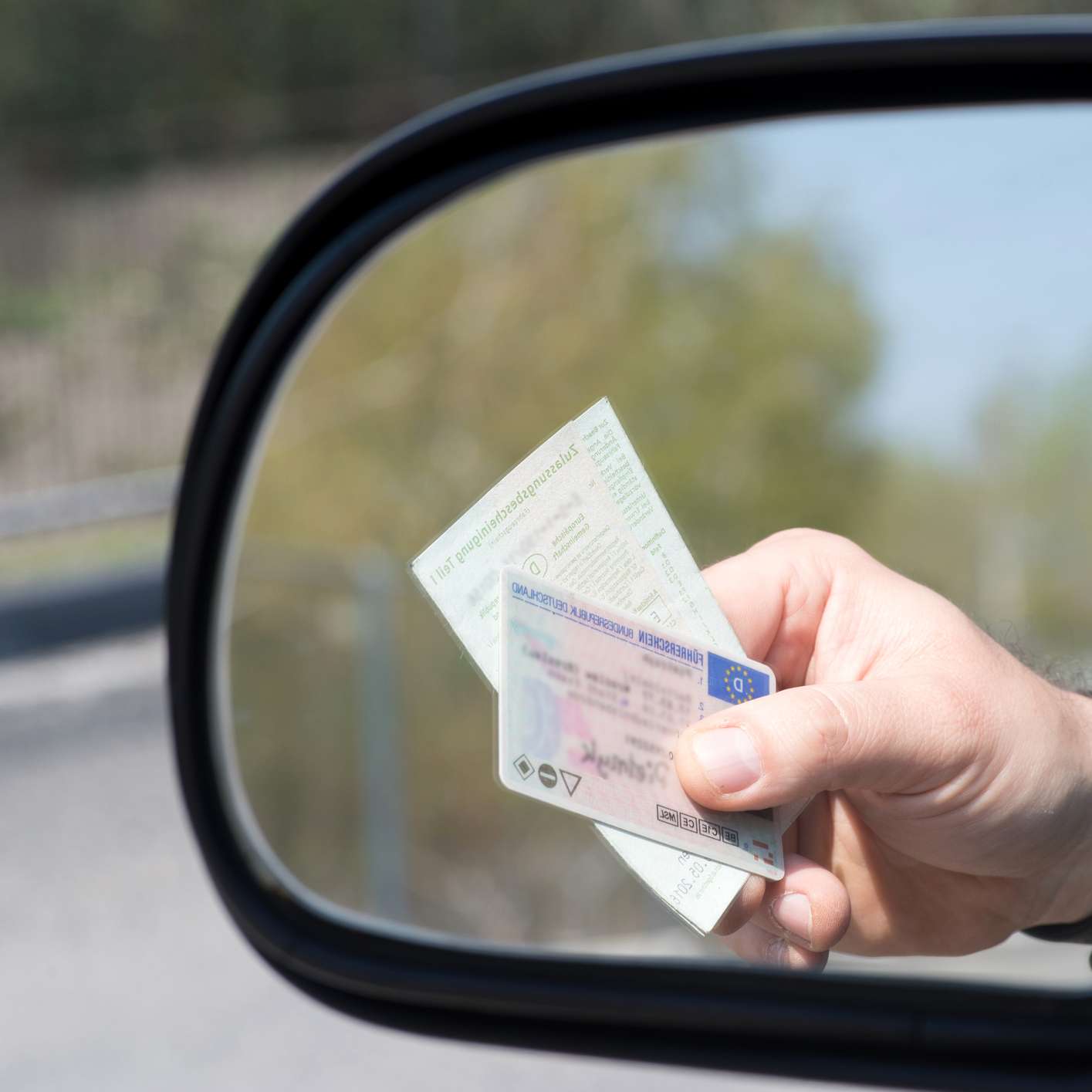 Autospiegel, in dem Hand des Fahrers zu sehen ist, der Fahrzeugschein und Führerschein hält 