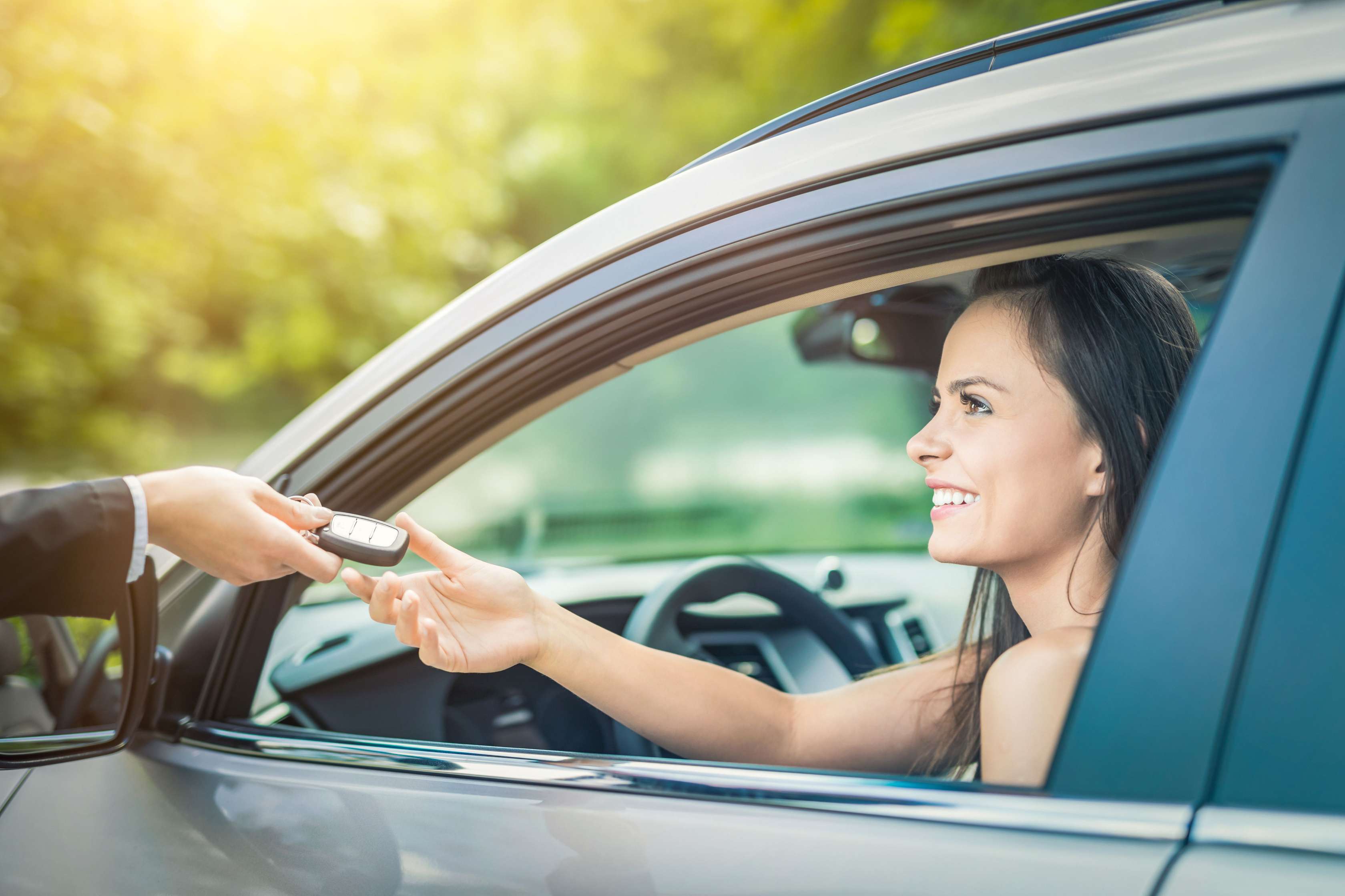 Hand übergibt junger Frau, die im Auto sitzt, durchs geöffnete Fahrerfenster den Autoschlüssel