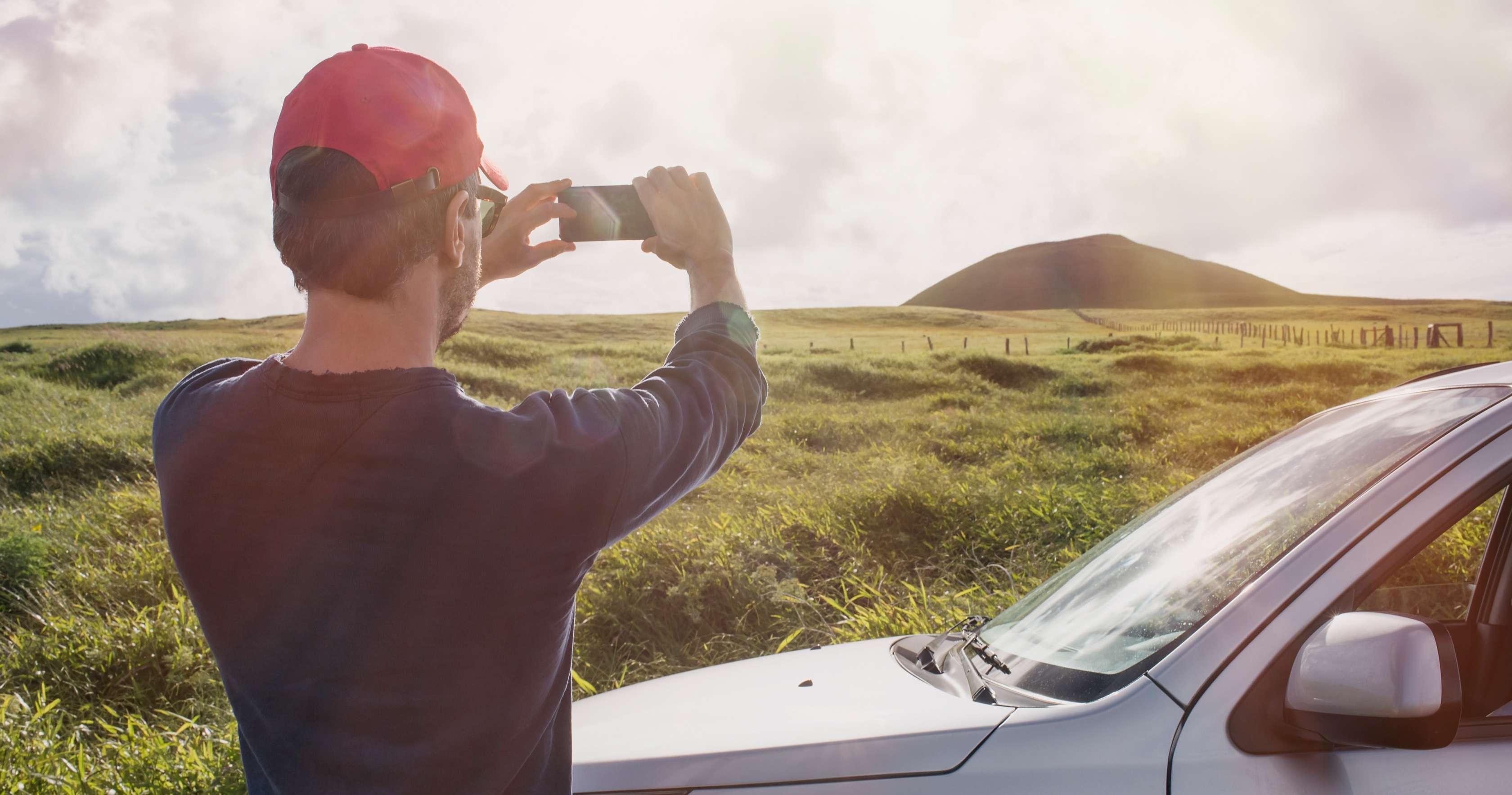 Mann mit Basecap steht neben seinem Auto und fotografiert eine Landschaft