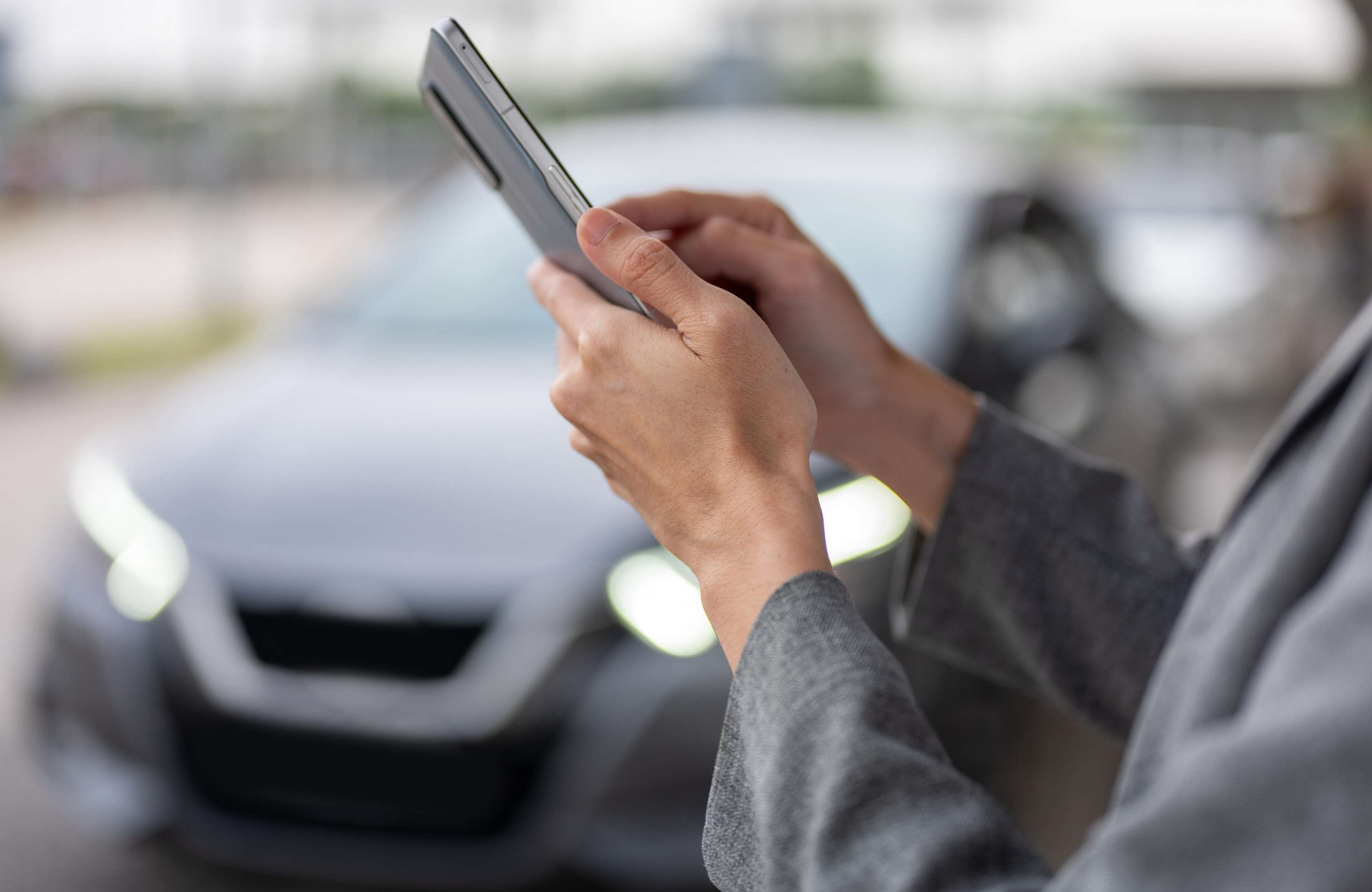 Kennzeichen verloren: Eine Frau steht vor einem Auto ohne nummernschild und tippt auf ihr Smartphone