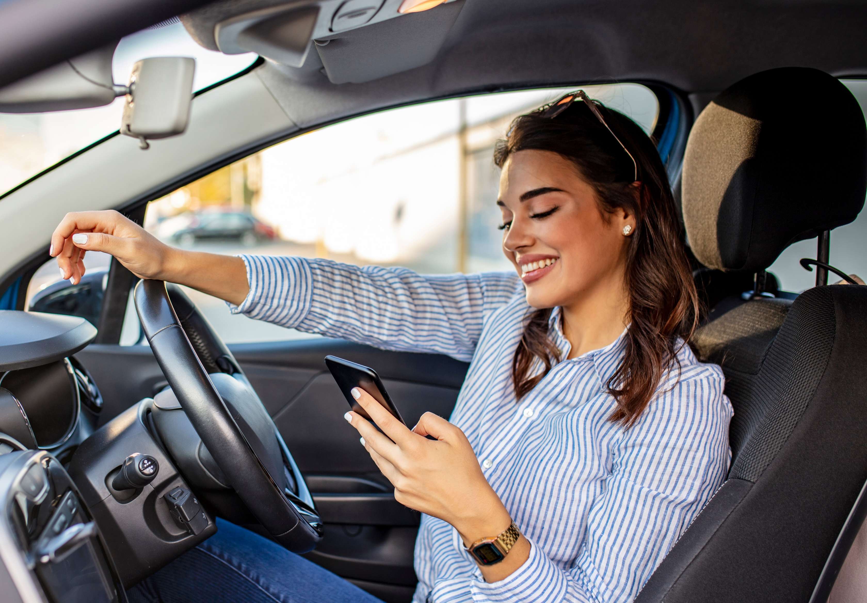 Kfz Versicherung Vergleich: Junge Frau sitzt am Steuer eines Pkw und blickt auf ihr Smartphone