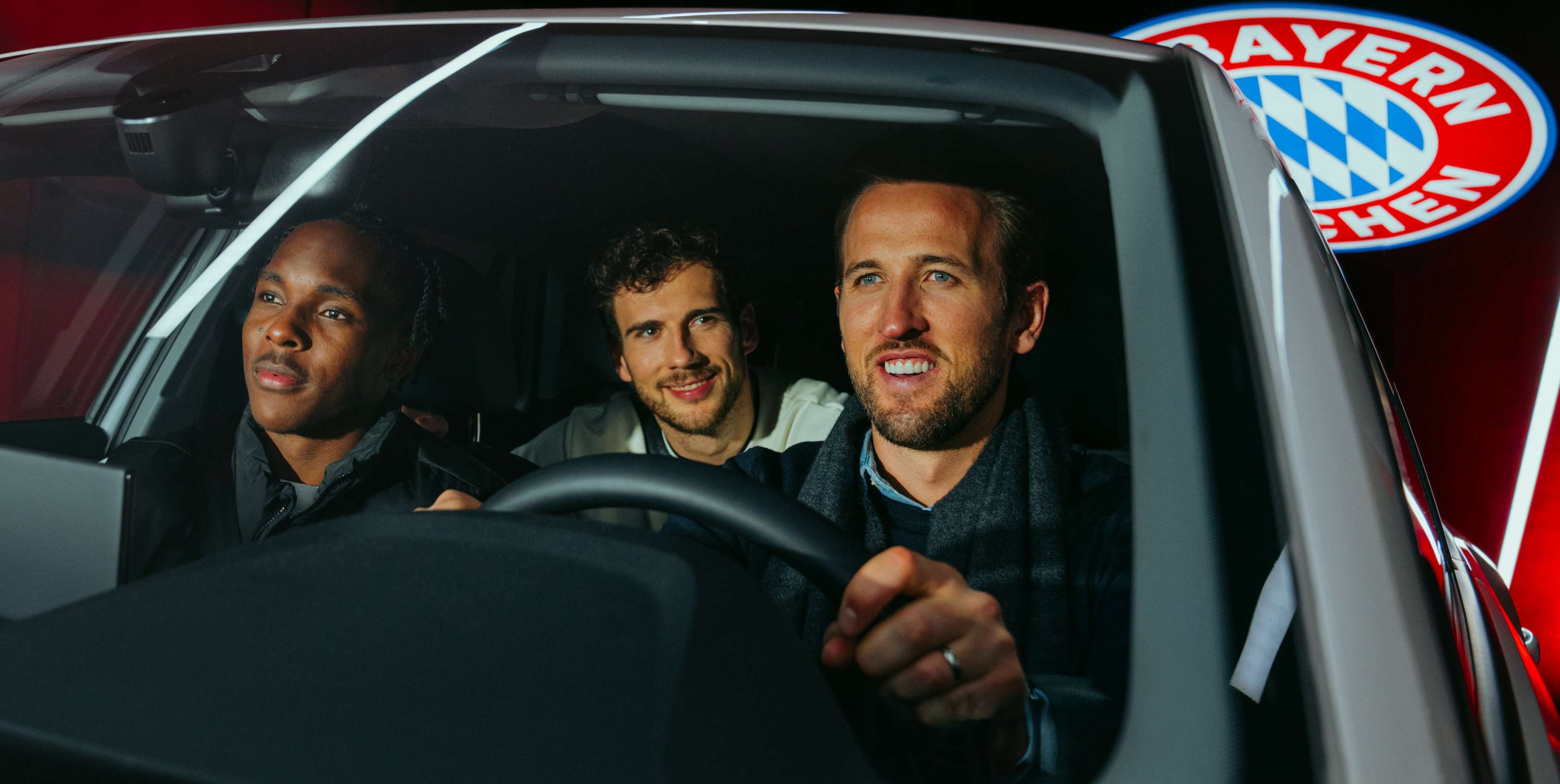 Kfz-Versicherung: Blick durch die Windschutzscheibe in ein Auto, an dessen Steuer Bayern-Spieler Harry Kane sitzt, Leon Goretzka ist auf dem Rücksitz, Mathys Tel auf dem Beifahrersitz 
