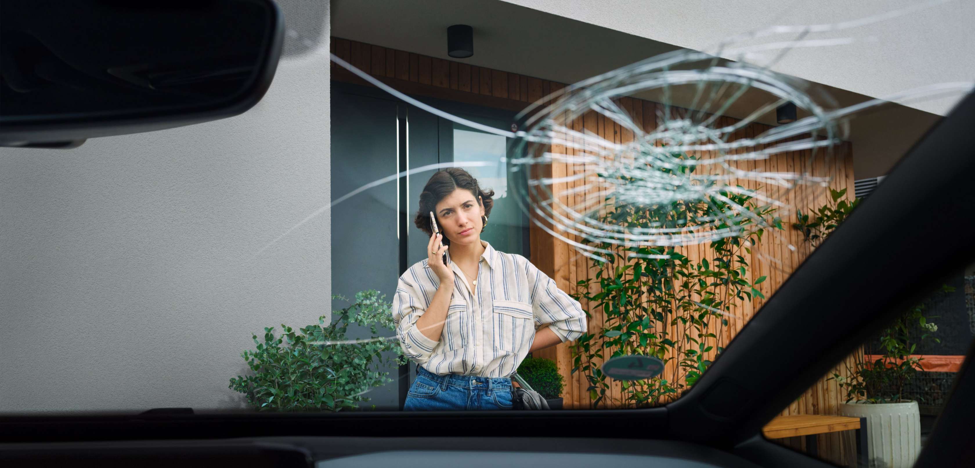 Junge Frau steht telefonierend vor ihrem Auto, das eine kaputte Windschutzscheibe ha