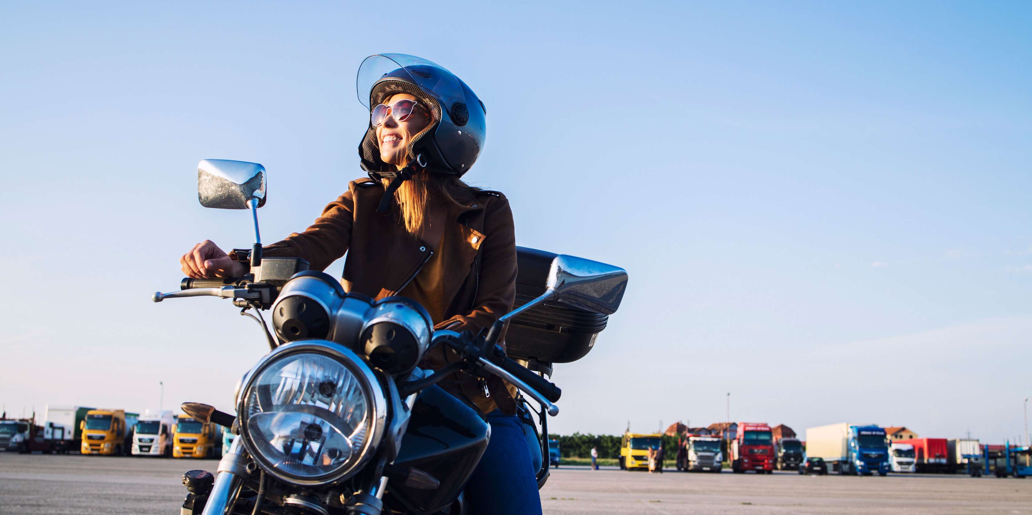 Kfz-Steuer Motorrad: Frau mit Lederjacke sitzt auf einem Motorrad auf einem Parkplatz in der Sonne