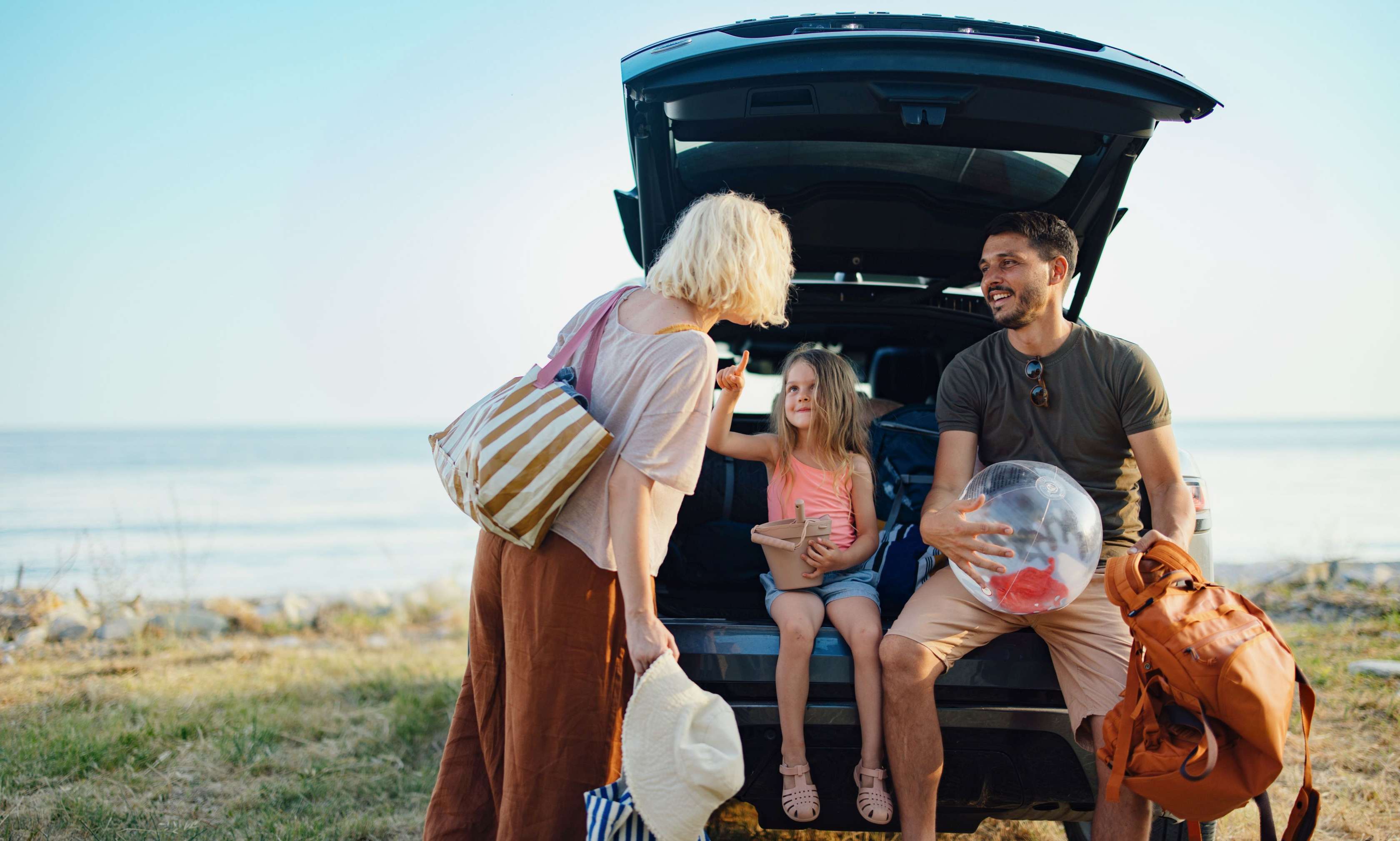 Familie sitzt entspannt und gut gelaunter Urlaubsstimmung im geöffneten Kofferraum eines Autos am Strand