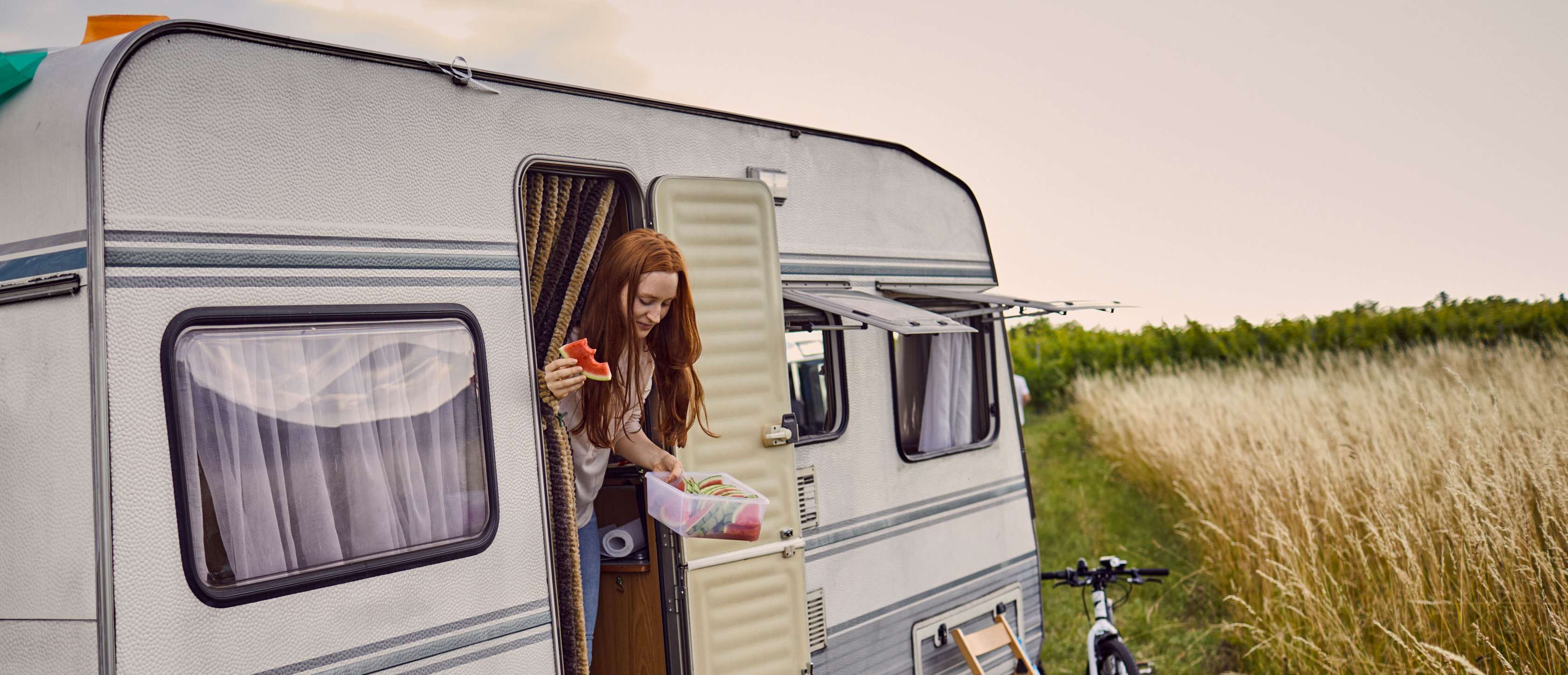 Wohnwagen Versicherung: Junge Frau steigt mit Wassermelone in der Hand aus Wohnwagen