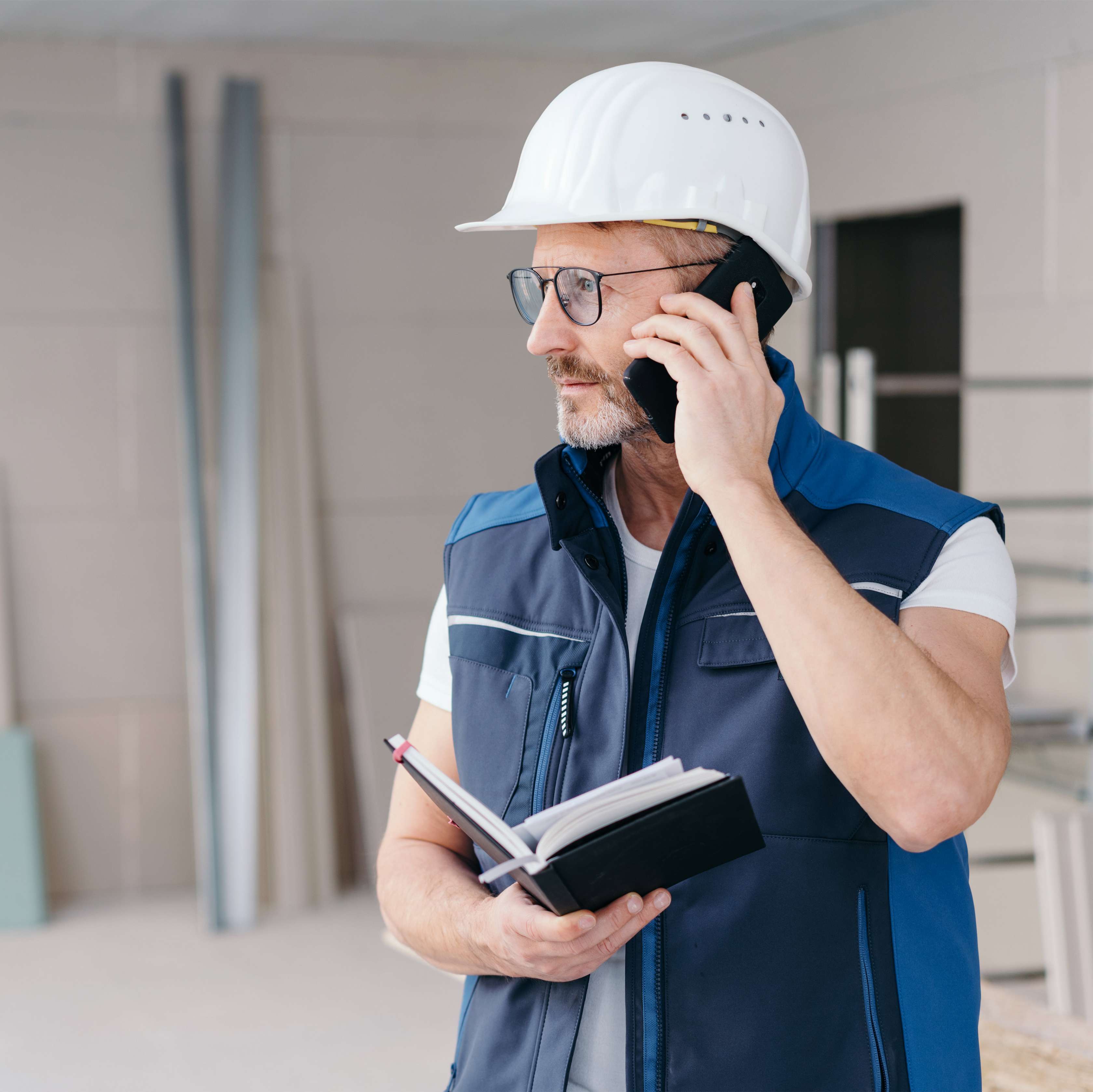 Ein Mann mit weißem Helm telefoniert auf einer Baustelle und hält dabei ein Notizbuch in der Hand