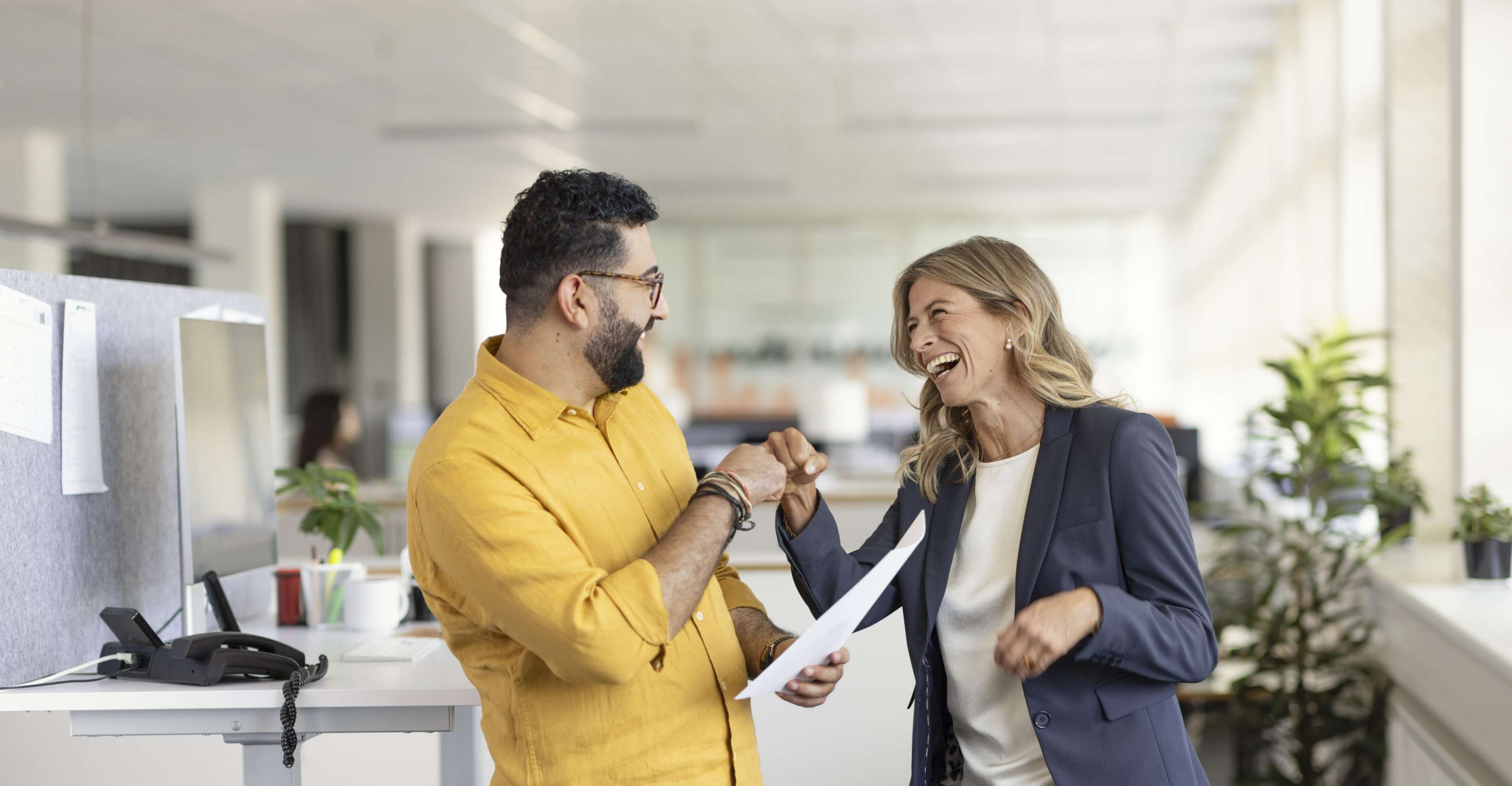 Allianz Betriebliche Altersversorgung für Unternehmen: Zwei Personen, ein Mann mit Brille und Bart in einem gelben Hemd sowie eine Frau mit langen blonden Haaren in einem dunkelblauen Blazer, lachen und geben sich dire Faust. Im Hintergrund ist ein Brüo mit Plfanzen zu sehen. 