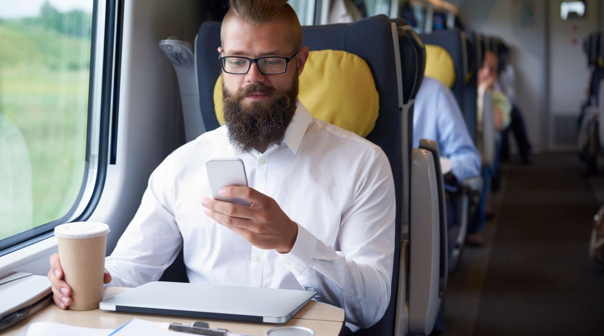 Allianz - Elektronikversicherung: Ein Mann sitzt in der Bahn, neben ihm Laptop und Kaffebecher