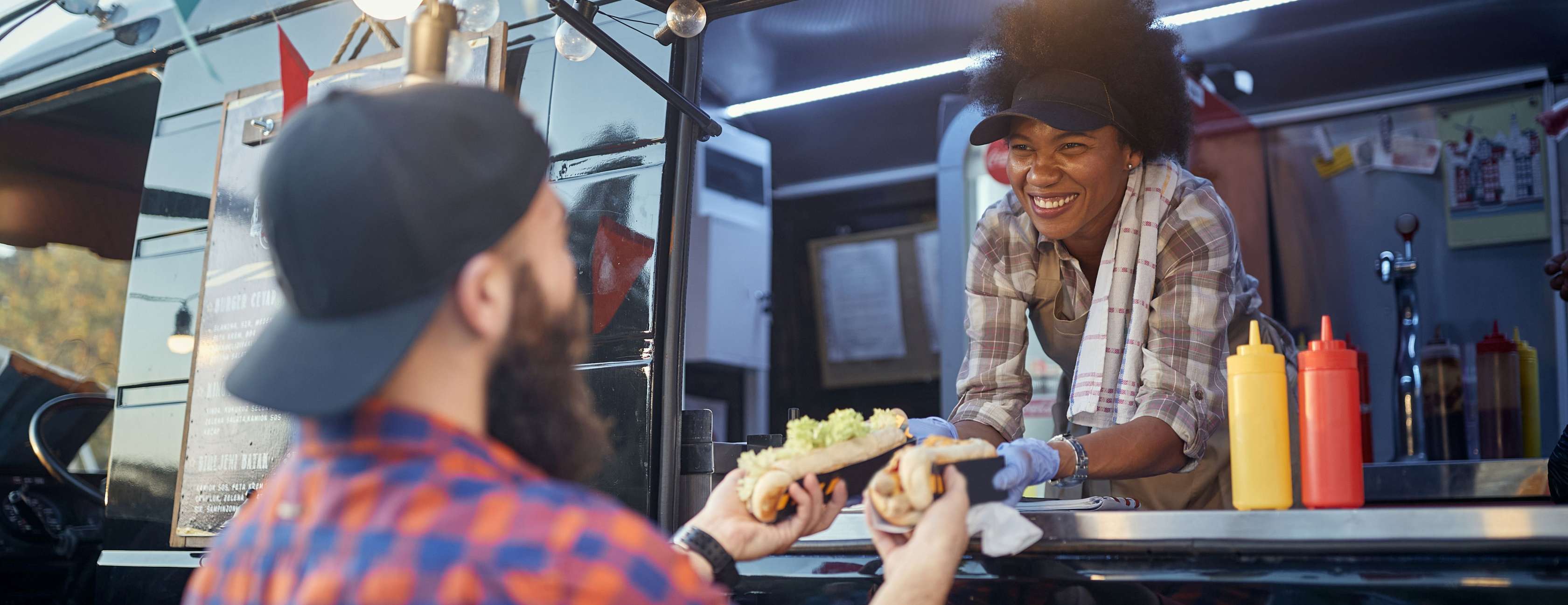 Eine Foodtruck-Betreiberin reicht einem Kunden lächelnd Hotdogs aus Ihrem Truck heraus.